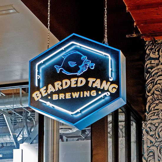 Hanging hexagonal neon sign for Bearded Tang Brewing inside Rodeo 39 Public Market, featuring a glowing blue neon border, illustrated tang fish, and bold gold lettering, suspended from chains against an exposed ceiling with a graphic mural column in the background.