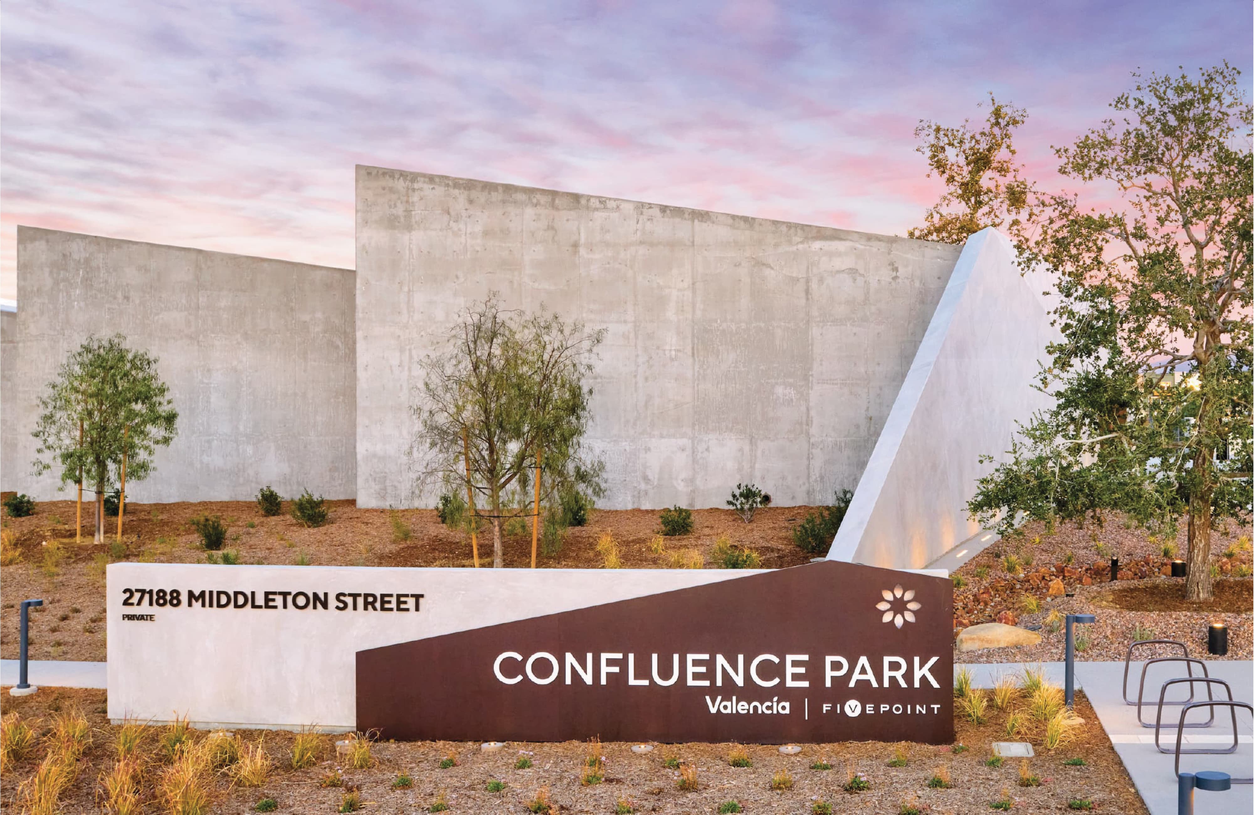 Angular monument sign reading 'Confluence Park, Valencia, FivePoint' with the address '27188 Middleton Street,' set against tall concrete walls and desert landscaping at dusk, with modern buildings visible beyond a wire bike rack.