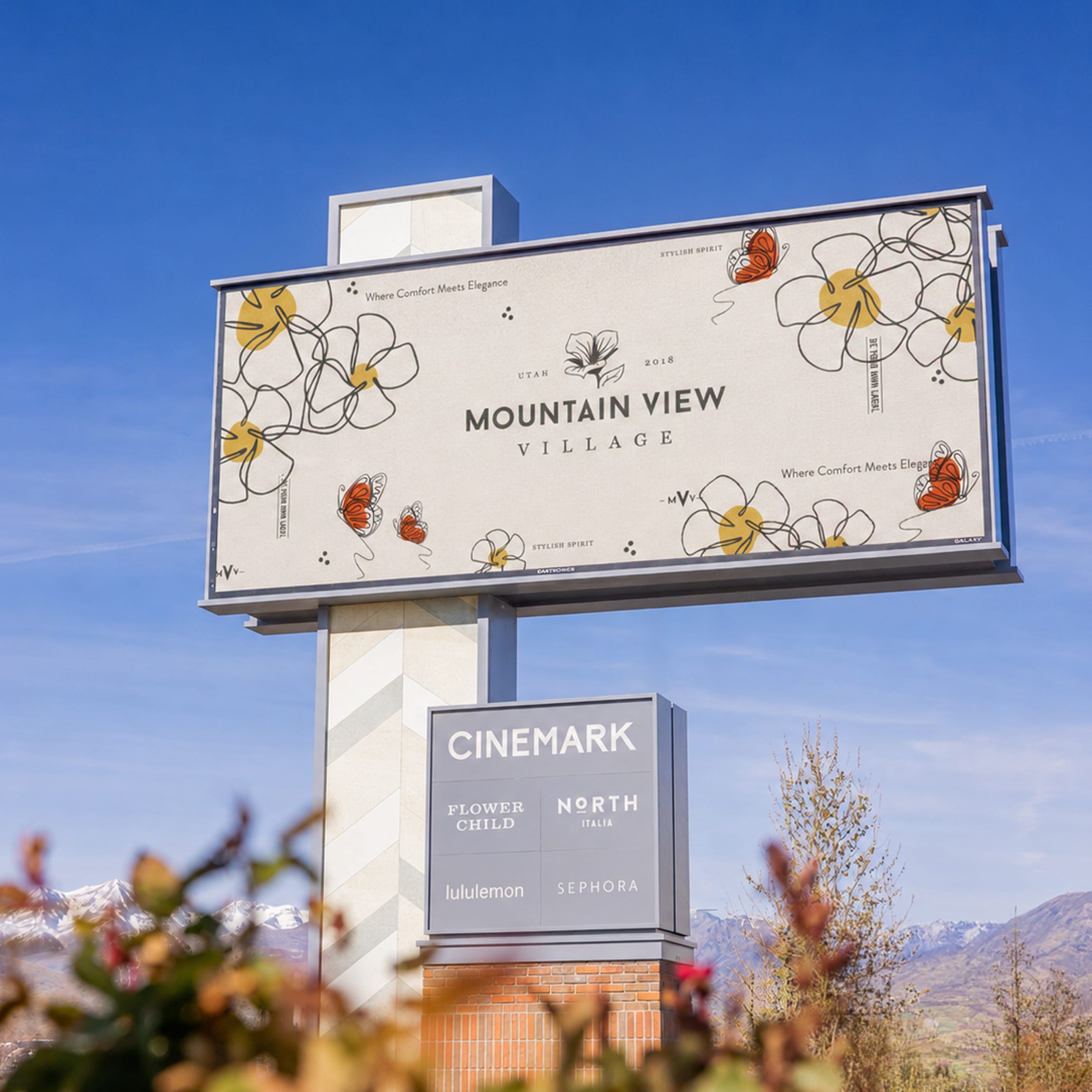 Elevated highway billboard with floral and butterfly branding for Mountain View Village above a tenant panel listing Cinemark, Flower Child, North Italia, lululemon, and Sephora, set against the Utah mountains in Riverton — branding, wayfinding, and environmental graphic design by RSM Design.