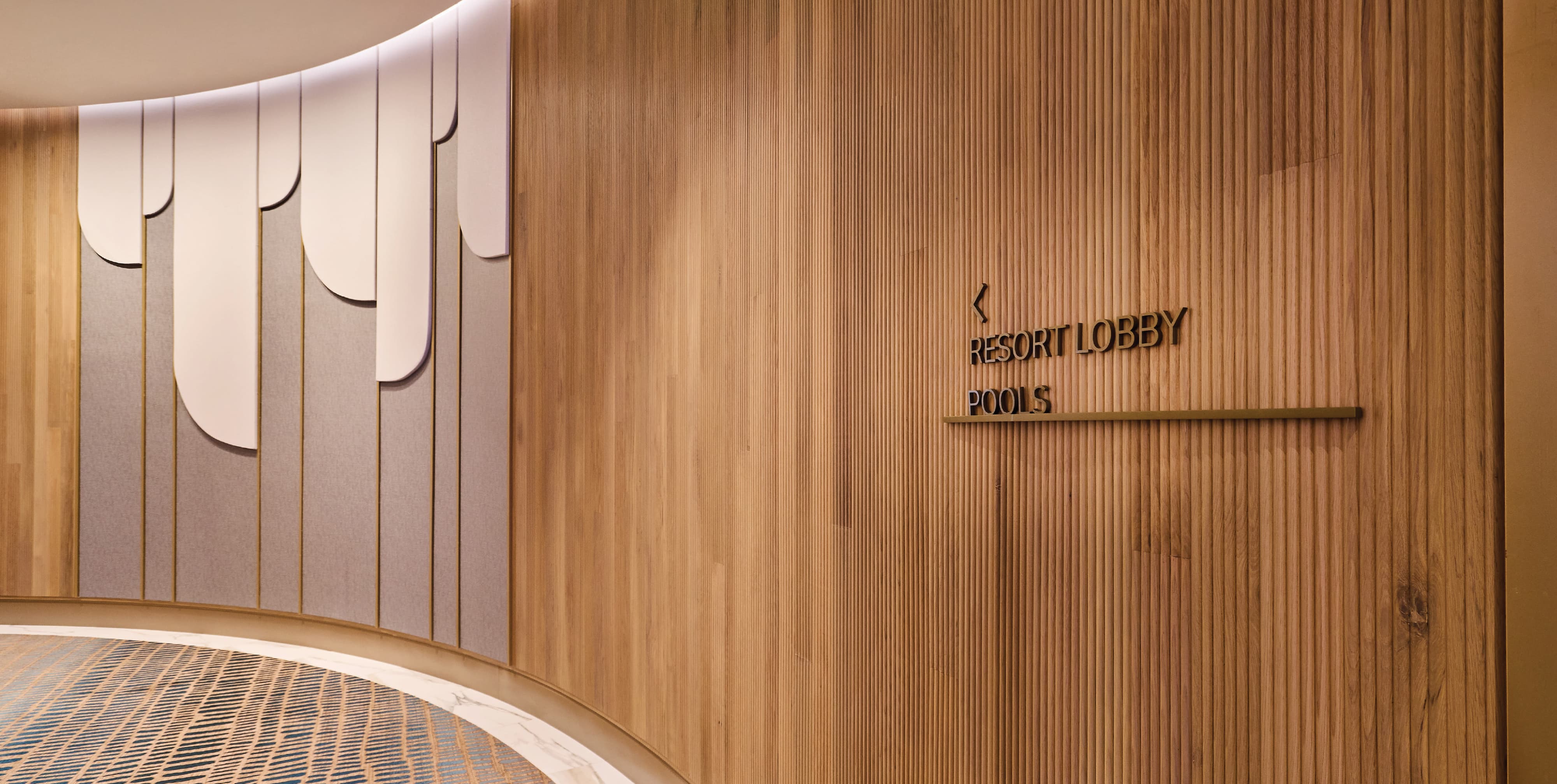 Interior corridor wayfinding signage directing guests to the resort lobby and pools, mounted on a warm wood slat wall alongside a curved decorative panel feature wall at Pier Sixty-Six luxury waterfront resort in Fort Lauderdale, Florida — RSM Design wayfinding and signage design, master sign programs, branded environments, and environmental graphic design for hospitality and waterfront destinations.