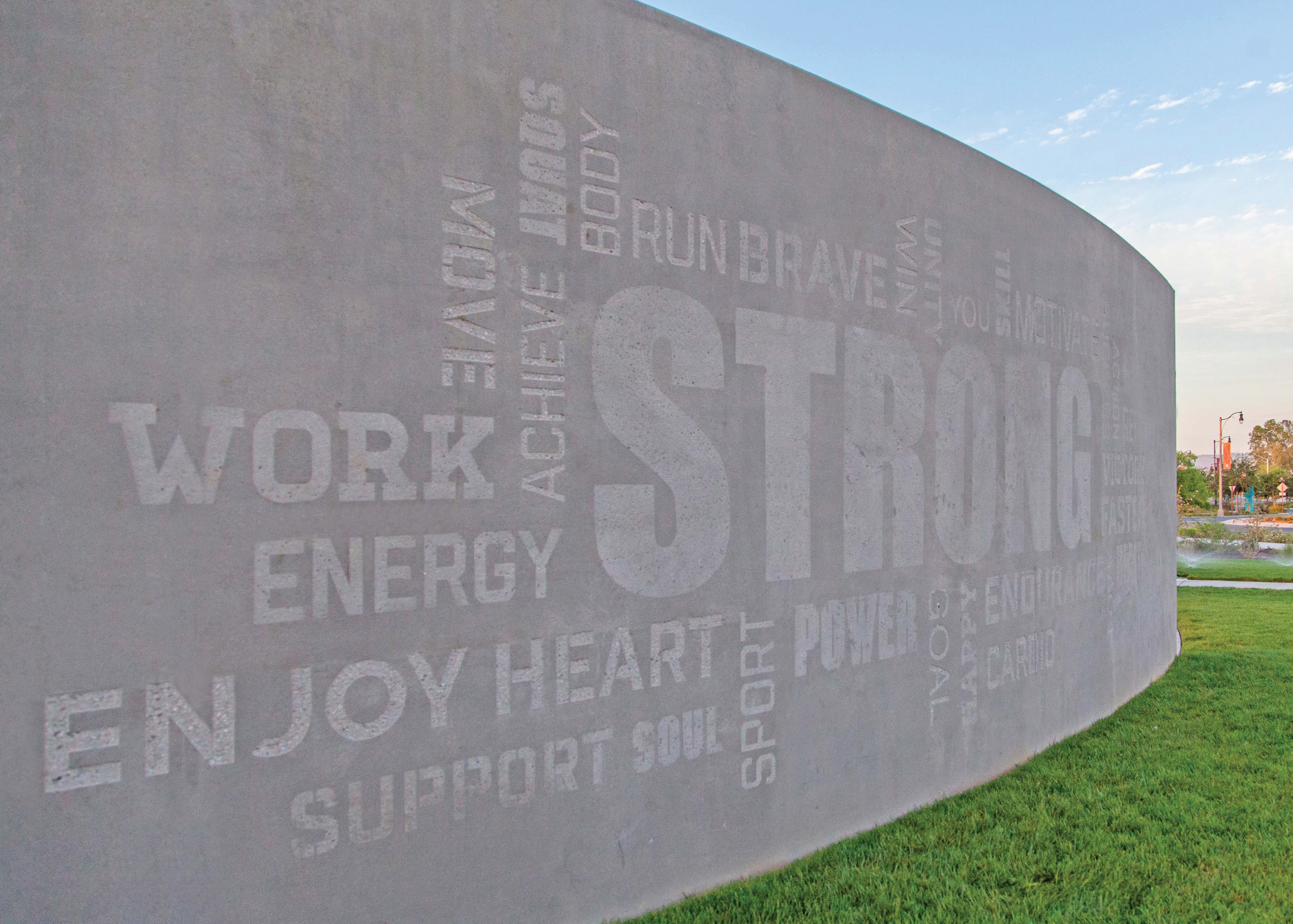 Curved concrete wall etched with a motivational word collage in varying sizes and orientations, featuring 'Strong' prominently among words like Work, Energy, Enjoy, Heart, Power, and Endurance, bordering a grassy lawn under a clear sky.