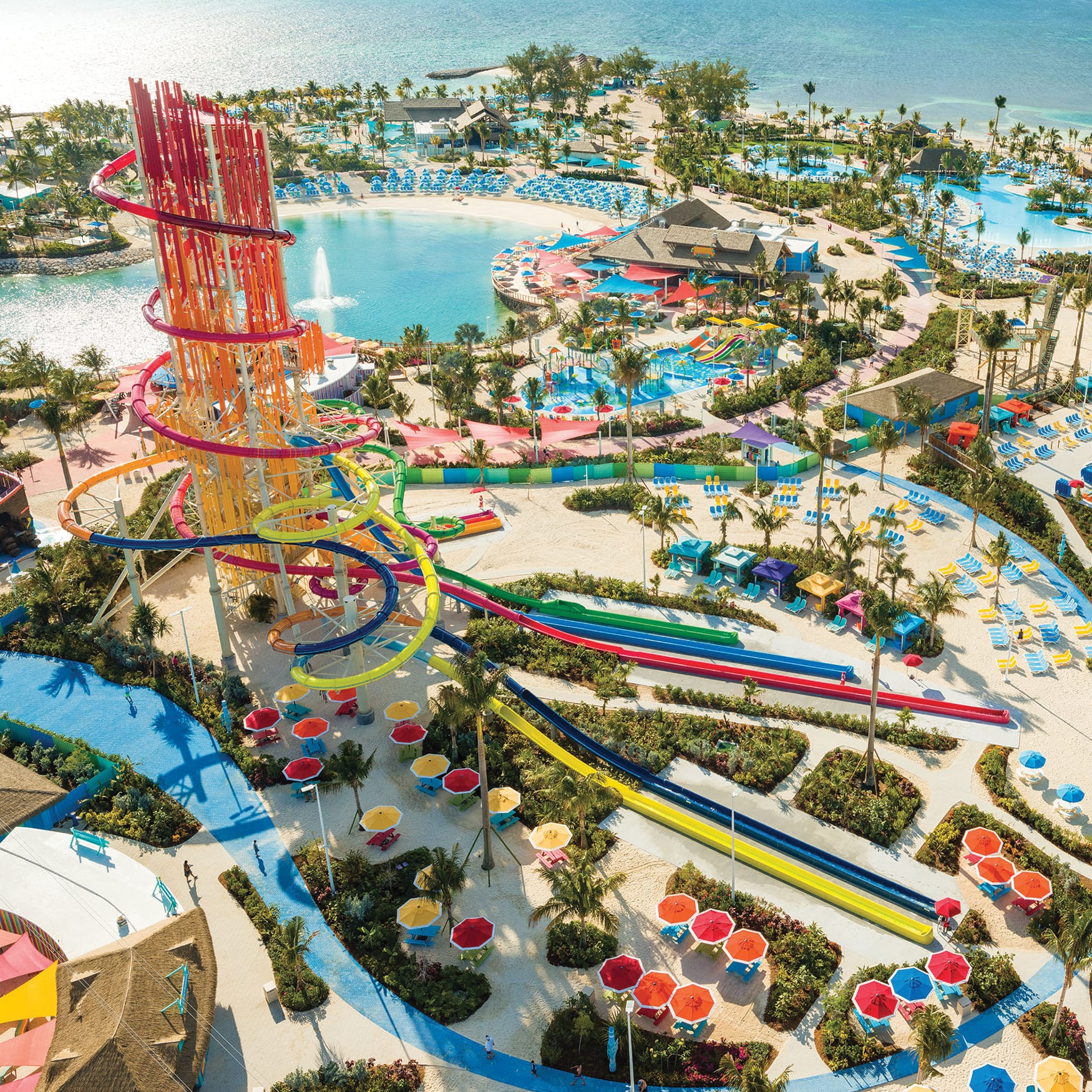 Aerial view of a tropical water park in the Bahamas featuring colorful twisting water slides, pools, sandy lounge areas, palm trees, and the turquoise ocean.