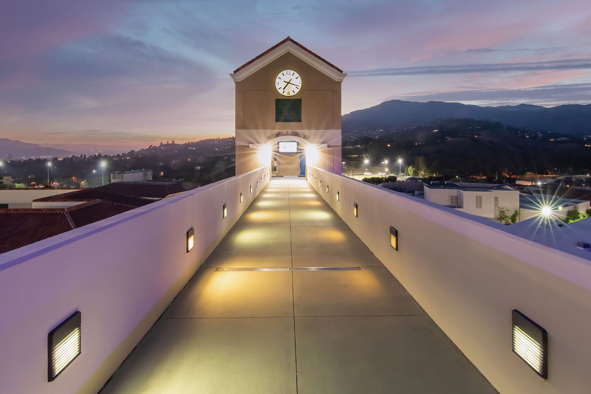 Illuminated pedestrian bridge leading to the Glendale Community College clock tower gateway at dusk, with warm pathway lighting, panoramic views of the San Gabriel Mountains, and a purple-hued sunset sky