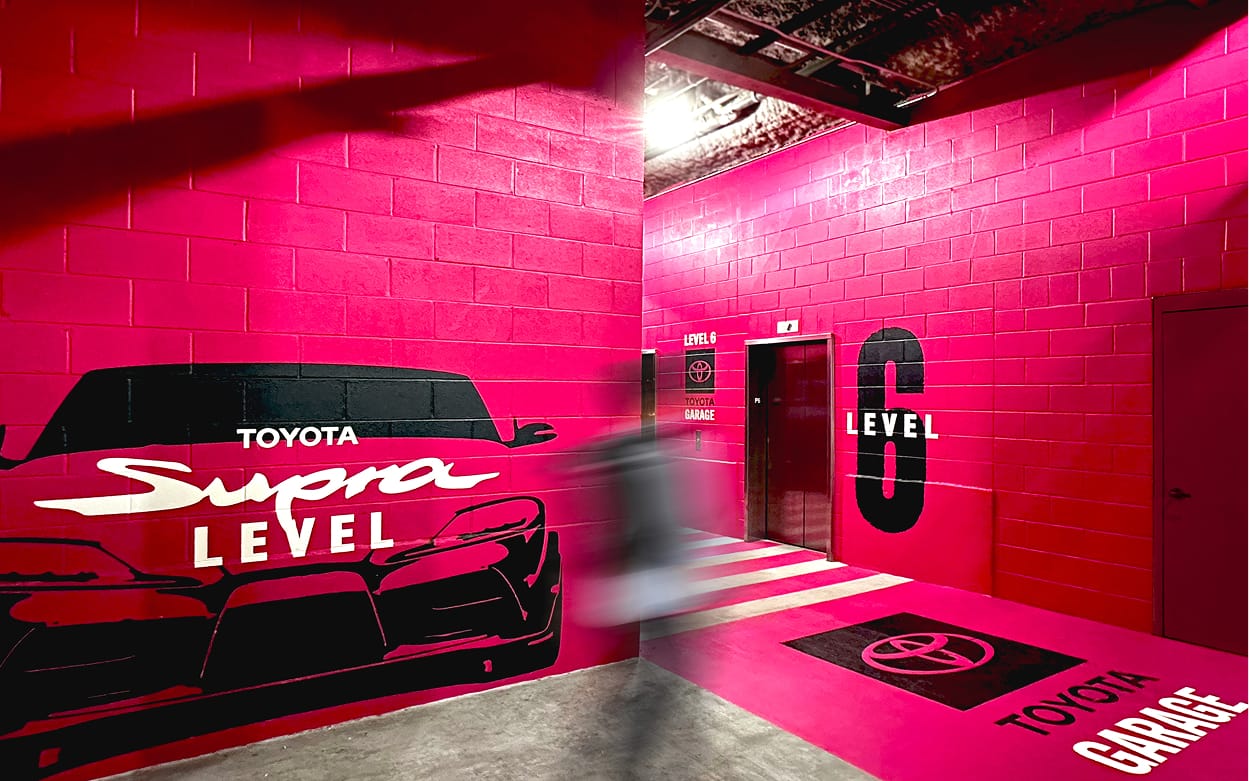 Toyota Supra branded Level 6 parking garage elevator lobby with immersive hot pink wall murals, large-scale vehicle graphics, level identification signage, and floor graphics at Toyota Music Factory in Irving, Texas — RSM Design experiential graphics, branded environments, wayfinding and signage design, specialty graphics, and master sign programs for parking garage and mixed-use destinations.