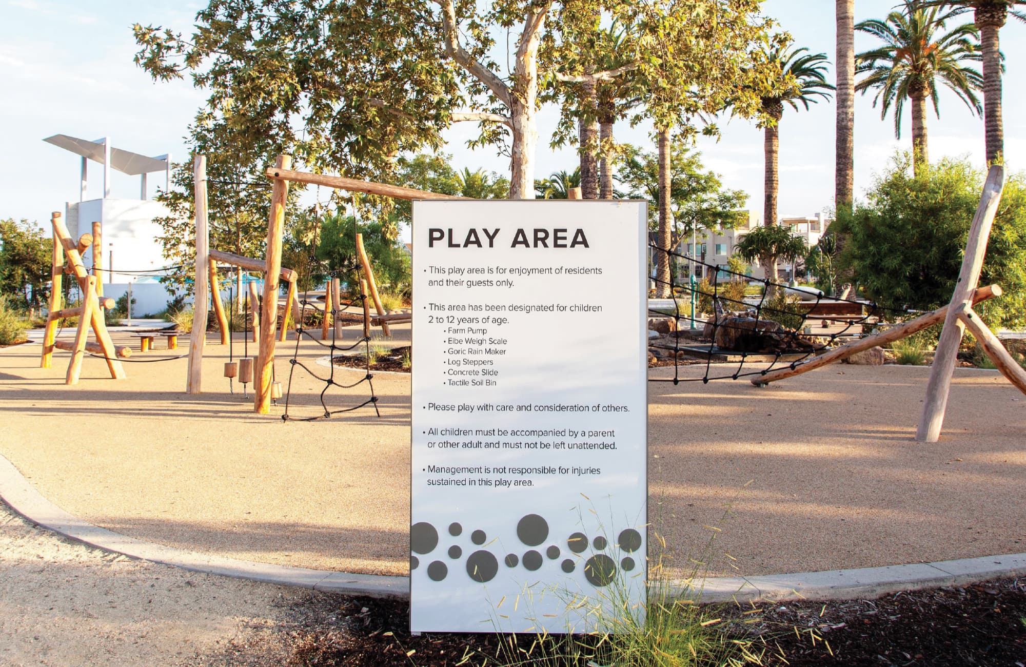 Play Area rules sign in front of a nature-themed playground with wooden climbing structures, rope nets, and a tactile soil bin, surrounded by palm trees and residential buildings.