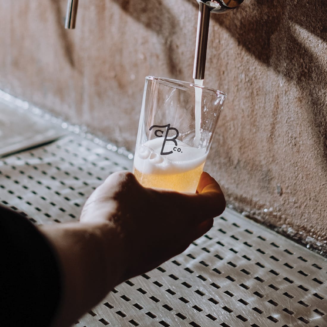 Hand holding a tasting glass with the CB Co. monogram while beer pours from a tap, brand identity design by RSM Design
