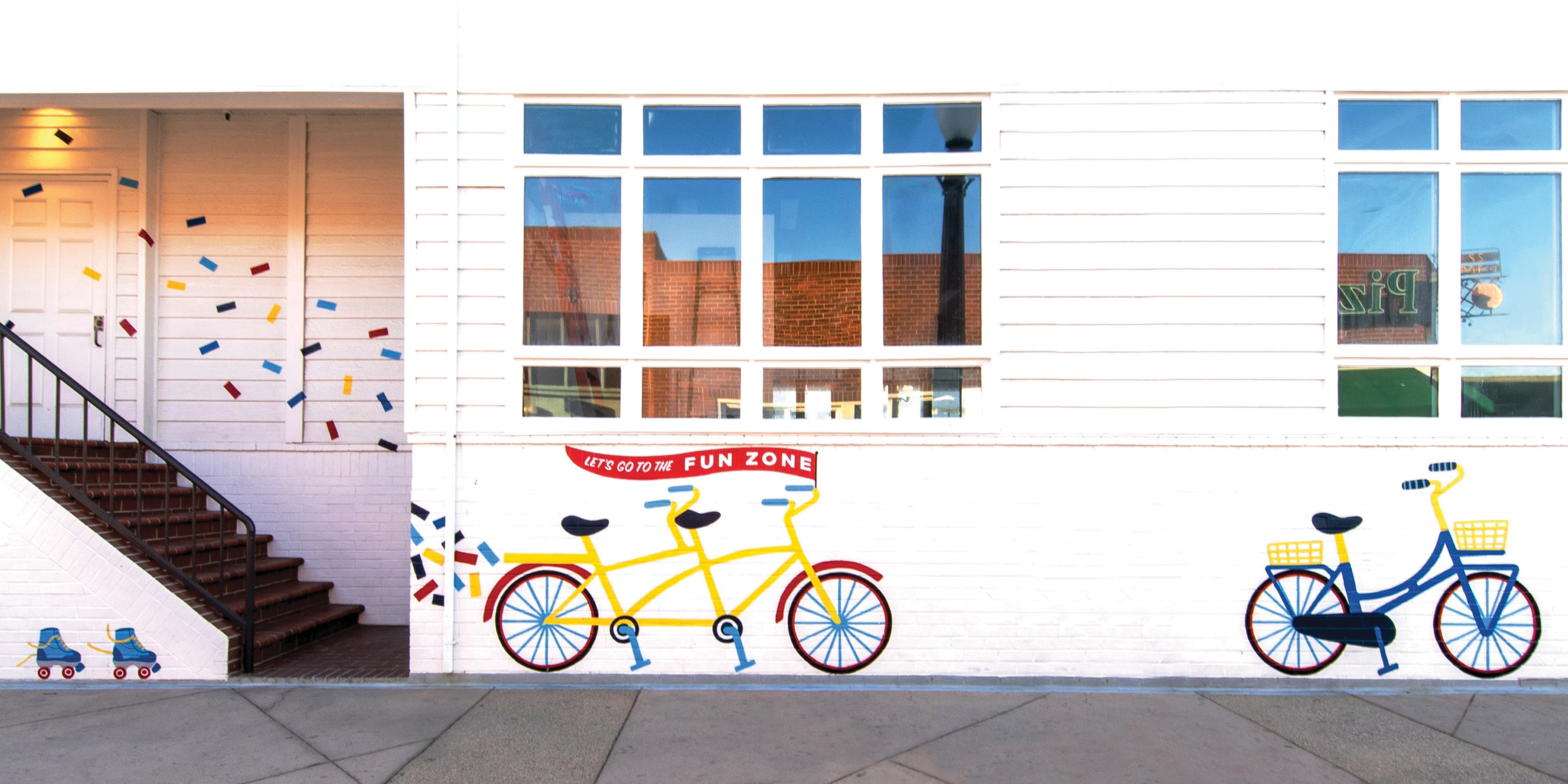 A wide exterior building mural at Balboa Fun Zone featuring a bold illustrated yellow tandem bicycle and a navy blue cruiser with a yellow basket, roller skates, colorful confetti shapes, and a red banner reading "Let's Go to the Fun Zone" painted across a white shiplap facade — showcasing RSM Design's art programs and public art and murals, experiential graphics and branded environments, and placemaking services within the waterfront and mixed-use markets
