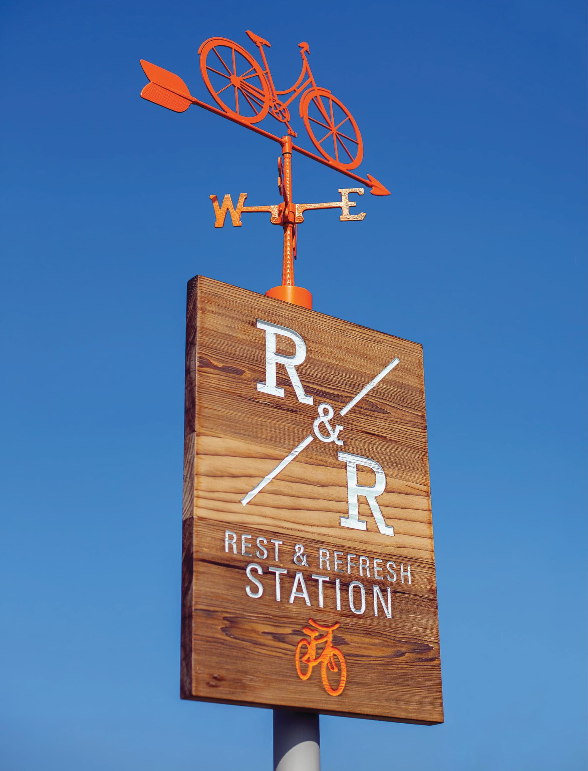 Wood sign reading 'R&R, Rest & Refresh Station' with a bicycle icon, topped by an orange bicycle-shaped weathervane, set against a clear blue sky.