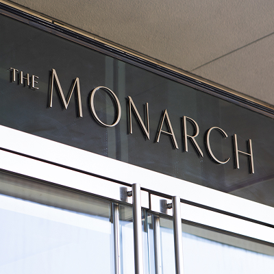 The Monarch dimensional lobby entrance signage — brushed metal letters on dark glass panel above glass entry doors, architectural interior signage by RSM Design, Frisco Texas