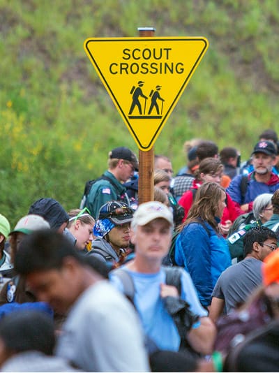 Yellow Scout Crossing pedestrian warning sign with hiker pictograms above crowd of Boy Scout Jamboree attendees at Summit Bechtel Reserve, custom outdoor wayfinding signage design by RSM Design