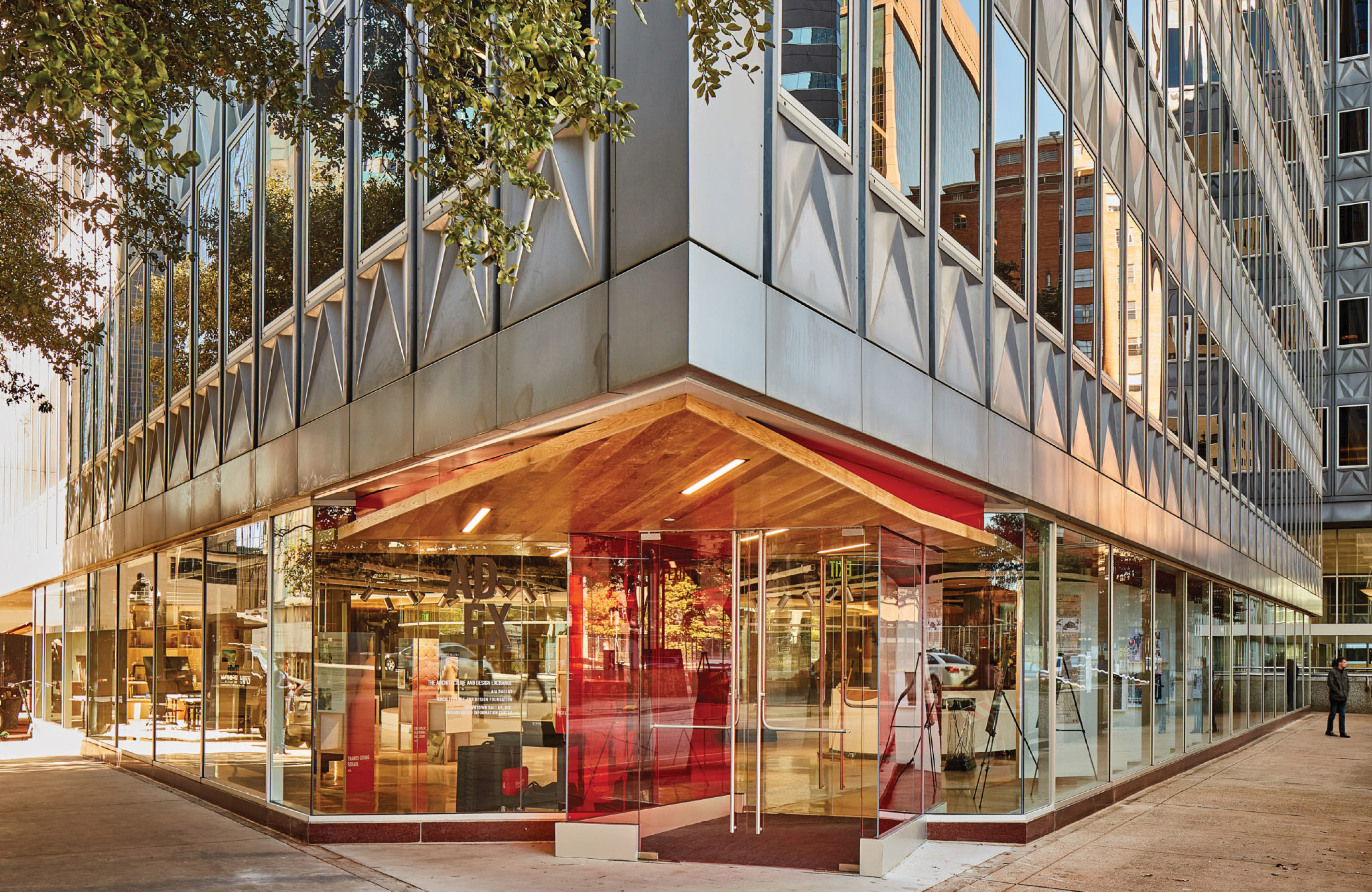 RSM Design exterior entry signage and environmental graphic design for the Architecture and Design Exchange in downtown Dallas, Texas, featuring window graphics and identity signage integrated into a dramatic glass curtain wall corner building with a warm wood canopy entry, red accent door panels, and a pedestrian on the sidewalk, showcasing how wayfinding and signage design, visual identity, and branded environments seamlessly merge with landmark modernist architecture to create a compelling, human-centered sense of arrival for cultural, workplace, and urban streetscape markets.