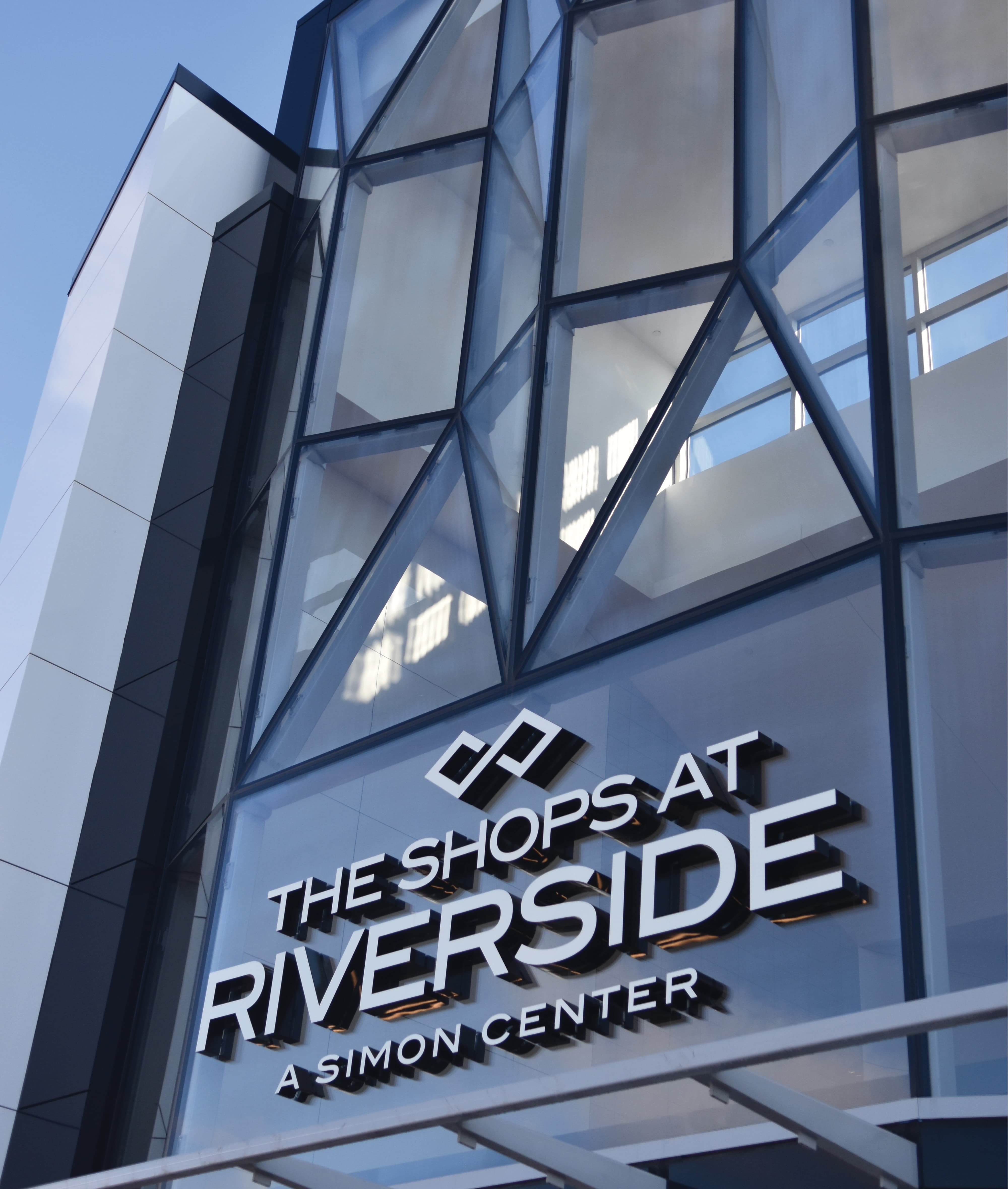 Branded exterior signage at The Shops at Riverside on a modern glass facade, showcasing environmental graphic design, placemaking, and identity design for retail, mixed-use, and commercial destination environments.