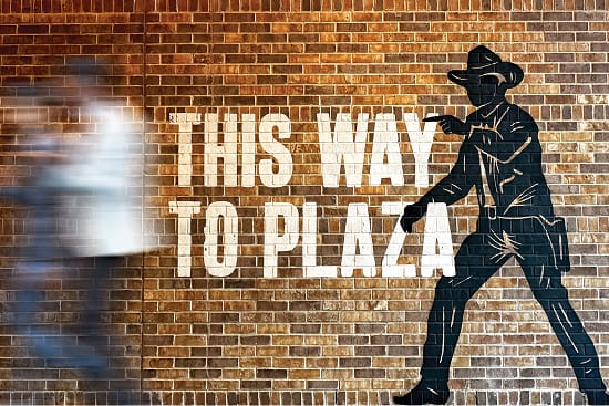 Pedestrian wayfinding mural on a brick wall featuring a cowboy silhouette and "This Way to Plaza" directional typography at Toyota Music Factory mixed-use entertainment district in Irving, Texas — RSM Design wayfinding and signage design, public art, murals, and experiential graphics for mixed-use, food and beverage, and streetscape destinations.
