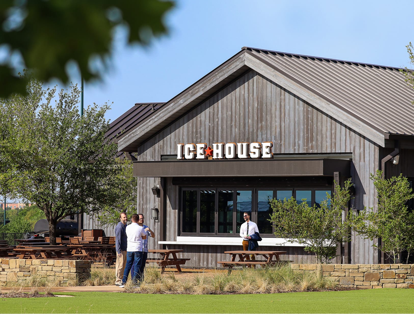 Exterior of Ice House bar and grill at PGA Frisco featuring rustic wood siding, metal roof, illuminated channel letter signage, outdoor picnic seating, and guests socializing on the lawn