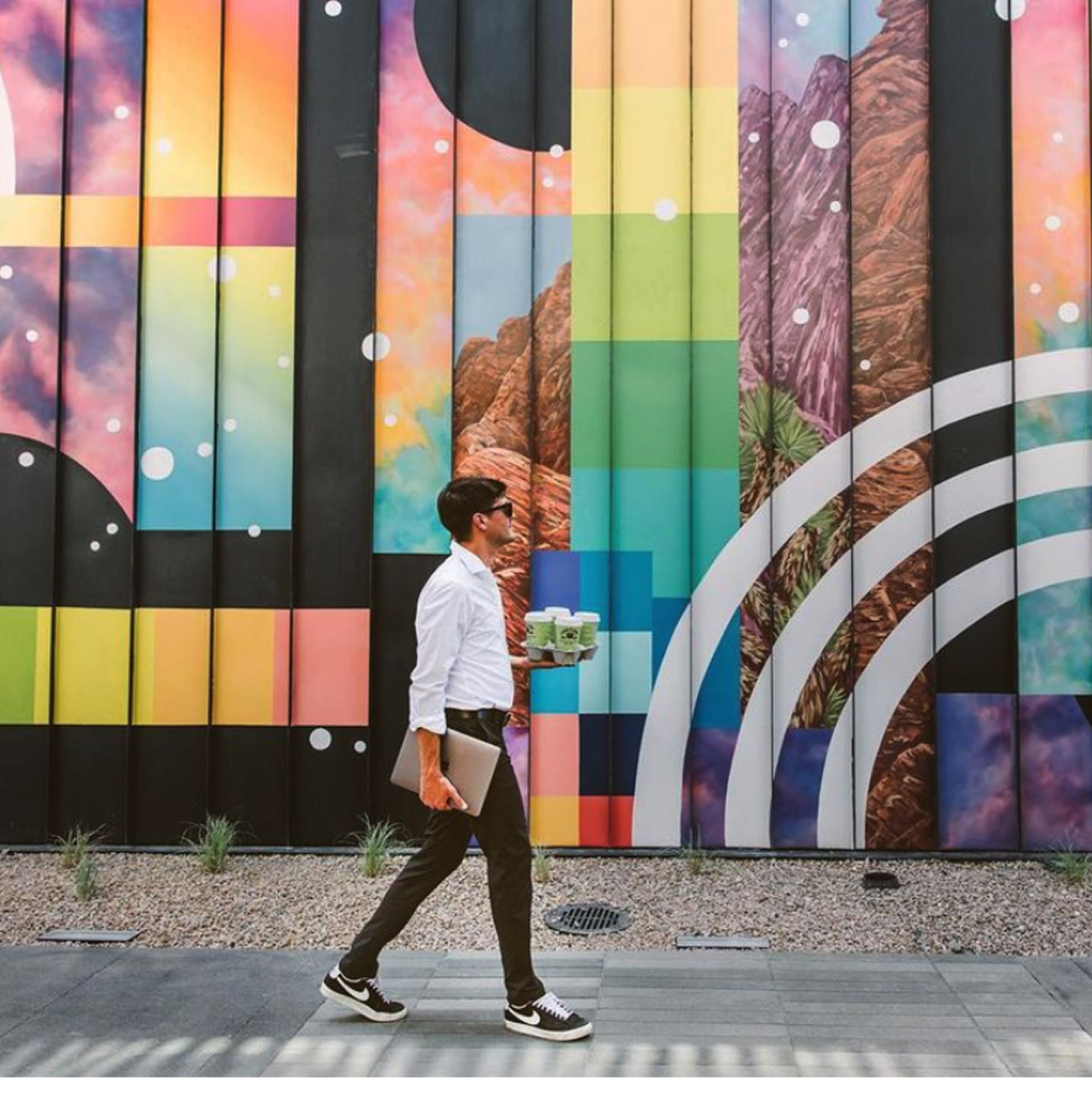 A person walks along a sidewalk in front of a large-scale, vibrant mural at Uncommons in Las Vegas. The mural combines colorful geometric rainbow stripes, cosmic imagery, and a realistic desert landscape featuring red rock formations and Joshua trees. The person carries a laptop under one arm and a tray of green drinks in the other.