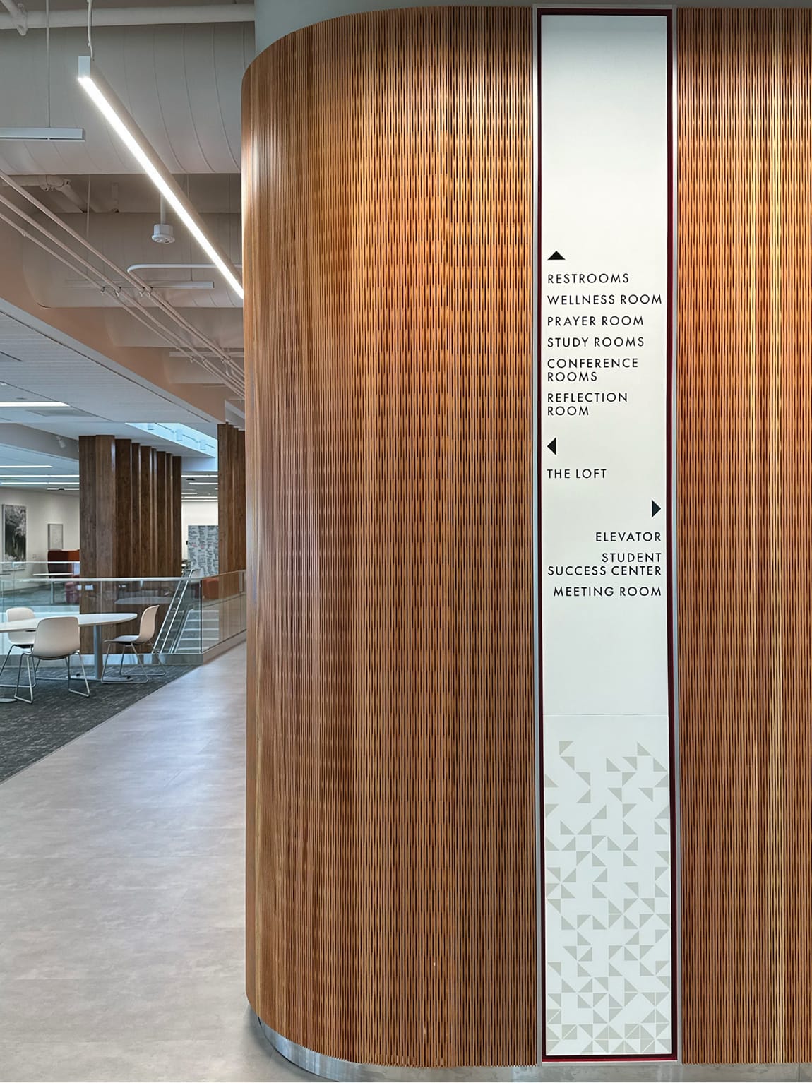 RSM Design Chapman University Rinker Campus Center Irvine — directional wayfinding signage panel mounted on curved wood slat feature wall, with geometric pattern detail and open student collaboration space beyond