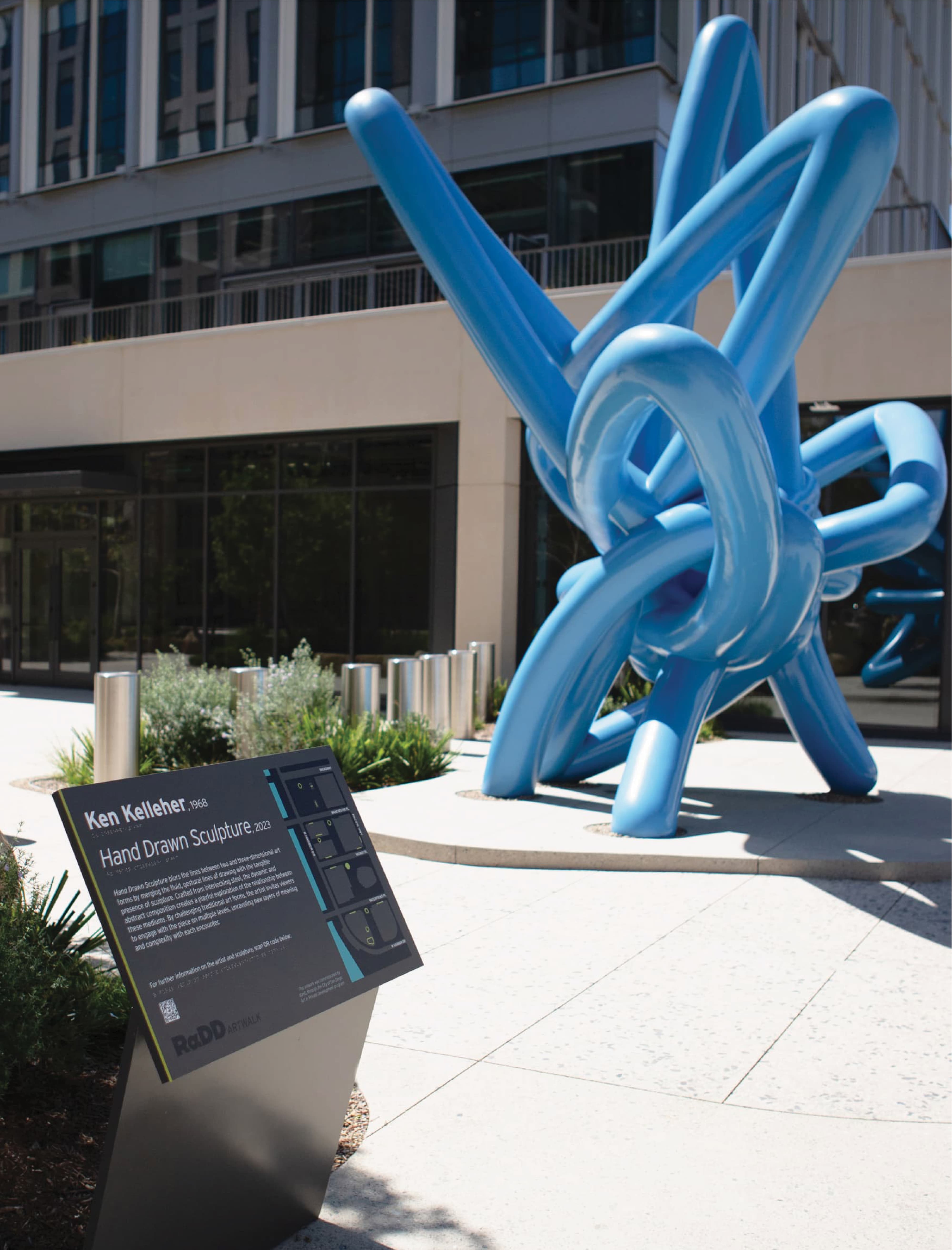 Large blue tubular sculpture of tangled, knotted loops titled 'Hand Drawn Sculpture' by Ken Kelleher, 2023, standing outside a modern building, with an interpretive plaque and map on a plinth in the foreground.