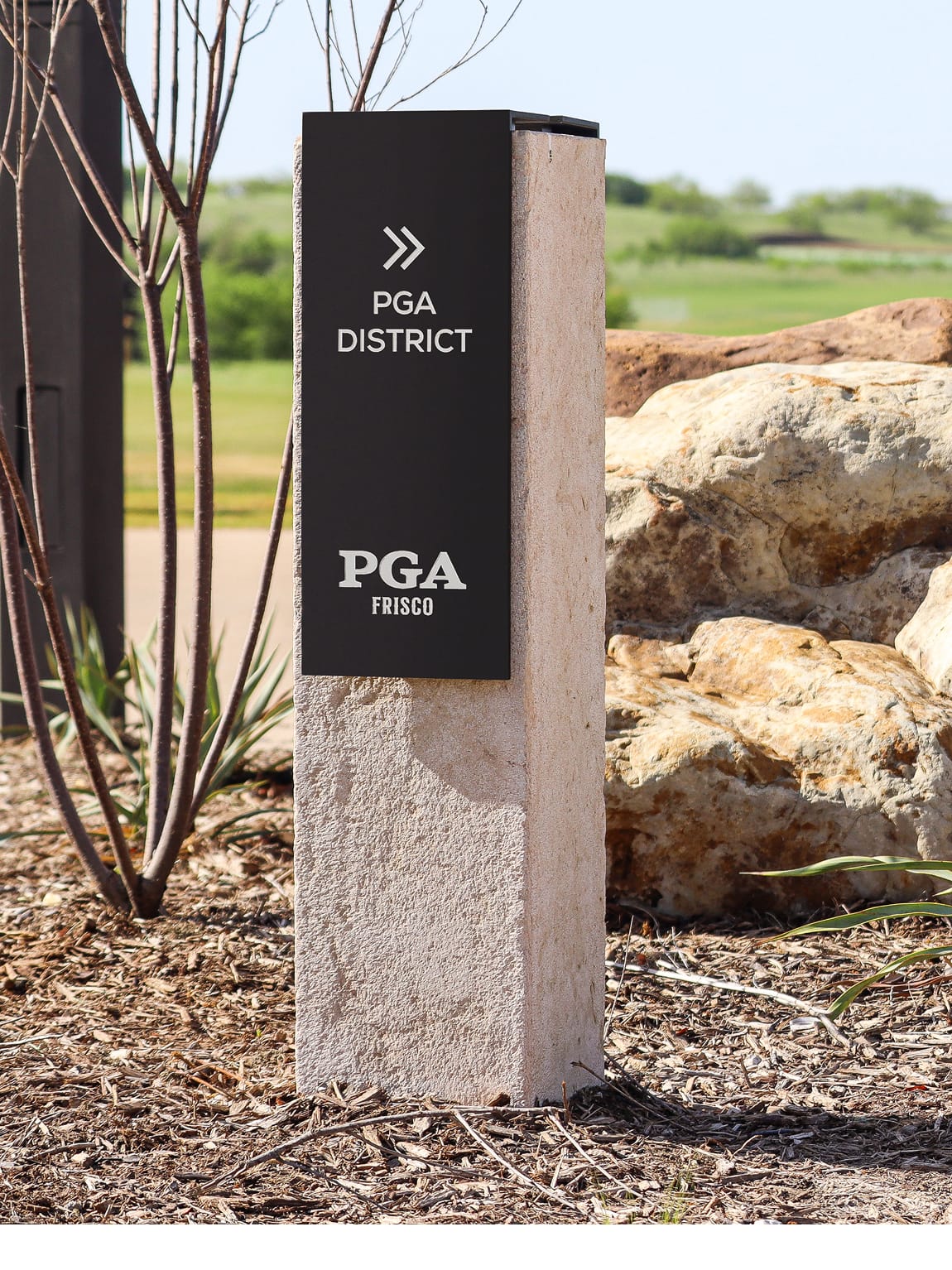 PGA Frisco District wayfinding bollard sign featuring black panel with double chevron arrow and PGA Frisco logo mounted on textured stone base, surrounded by native landscaping and boulders