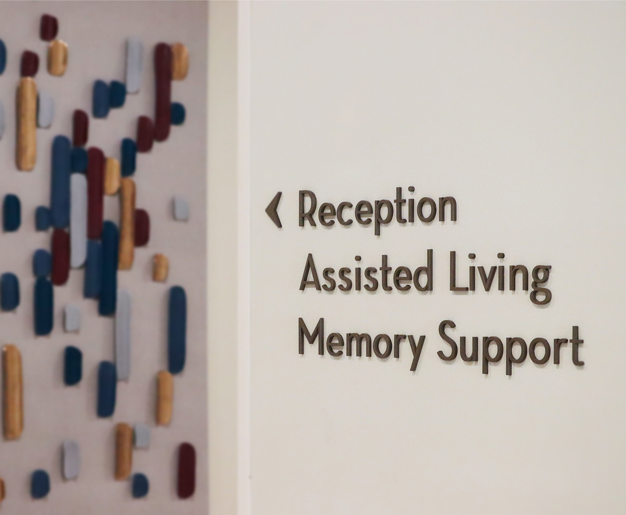 Wall directional sign reading 'Reception, Assisted Living, Memory Support' with a left-pointing arrow, beside a textured art panel of colorful vertical tabs in navy, burgundy, gold, and gray.