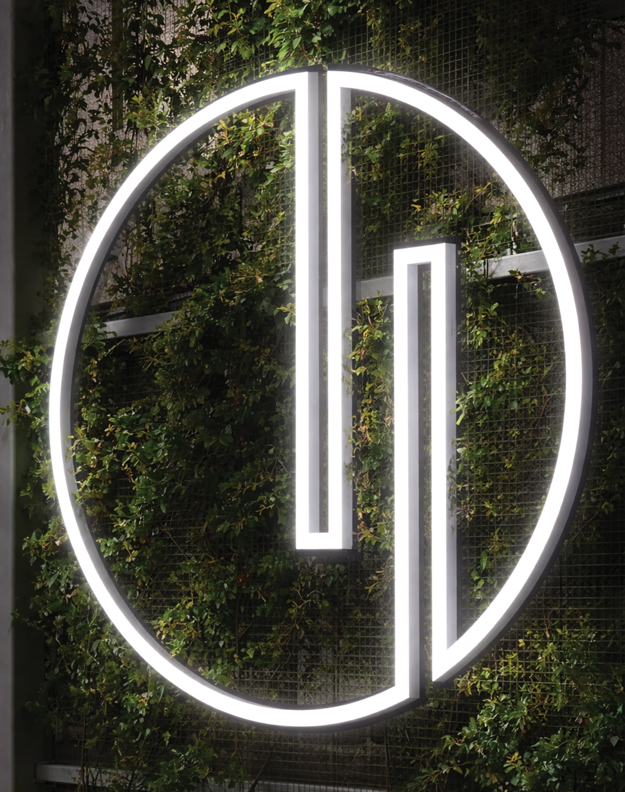 Illuminated white neon circular logo mark mounted against a lush green living wall, showcasing experiential graphics and branded environment design for a mixed-use workplace development by RSM Design.