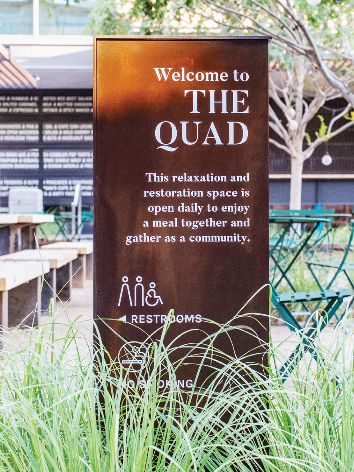 A Corten steel freestanding sign in an outdoor seating area reads "Welcome to The Quad" with the description: "This relaxation and restoration space is open daily to enjoy a meal together and gather as a community." Below the text are icons and directional information for restrooms and a no smoking notice. Ornamental grasses frame the base of the sign, with green bistro tables and trees visible in the background.
