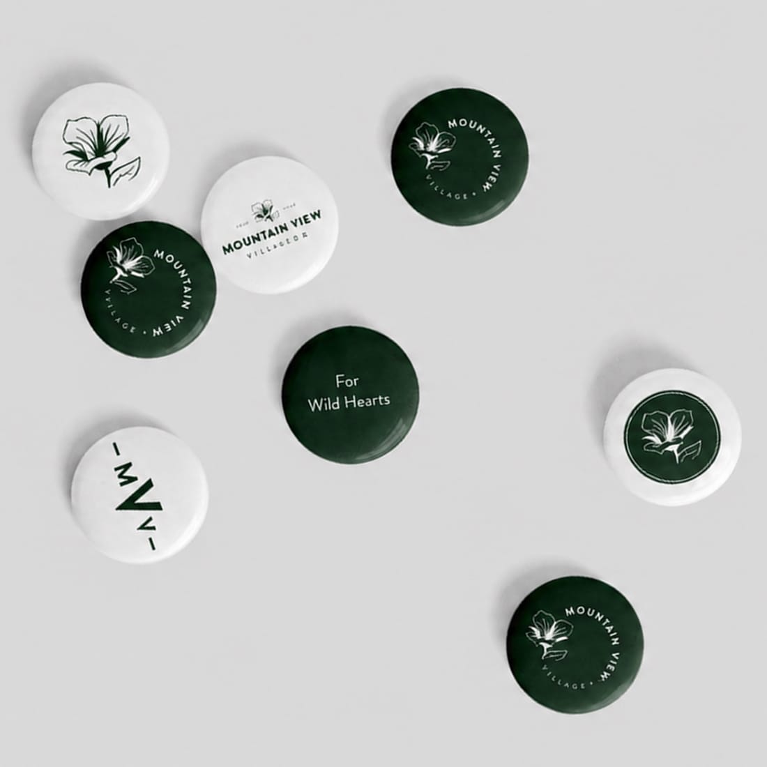Set of eight round pin badges in green and white featuring the Mountain View Village logo, sego lily emblem, "MVV" monogram, and "For Wild Hearts" tagline — branding and environmental graphic design by RSM Design.