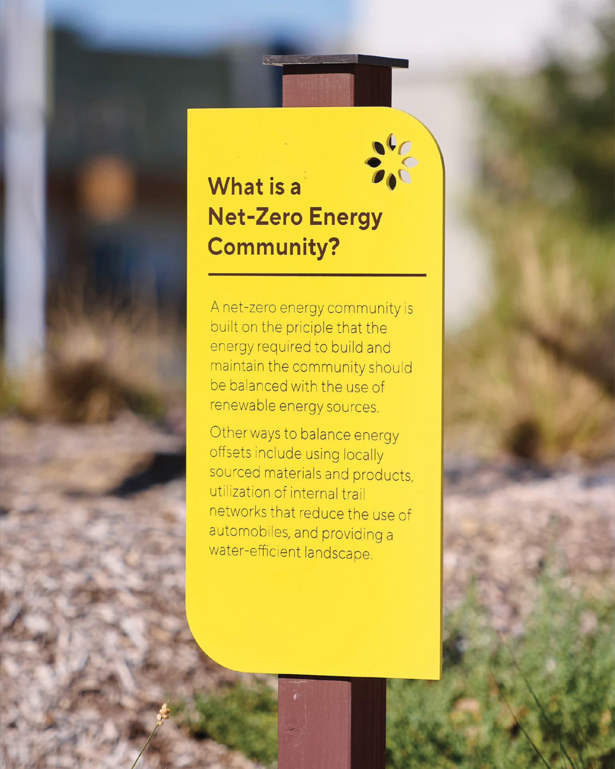 Yellow educational sign titled 'What is a Net-Zero Energy Community?' with a decorative flower cutout, mounted on a wooden post amid blurred desert landscaping.