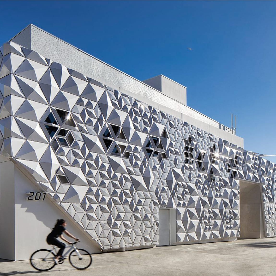 Full exterior view of a modern two-story building at 201 featuring an award-winning three-dimensional white geometric triangular cladding system, diamond-shaped windows integrated into the facade, and a motion-blurred cyclist passing in front against a vivid blue sky