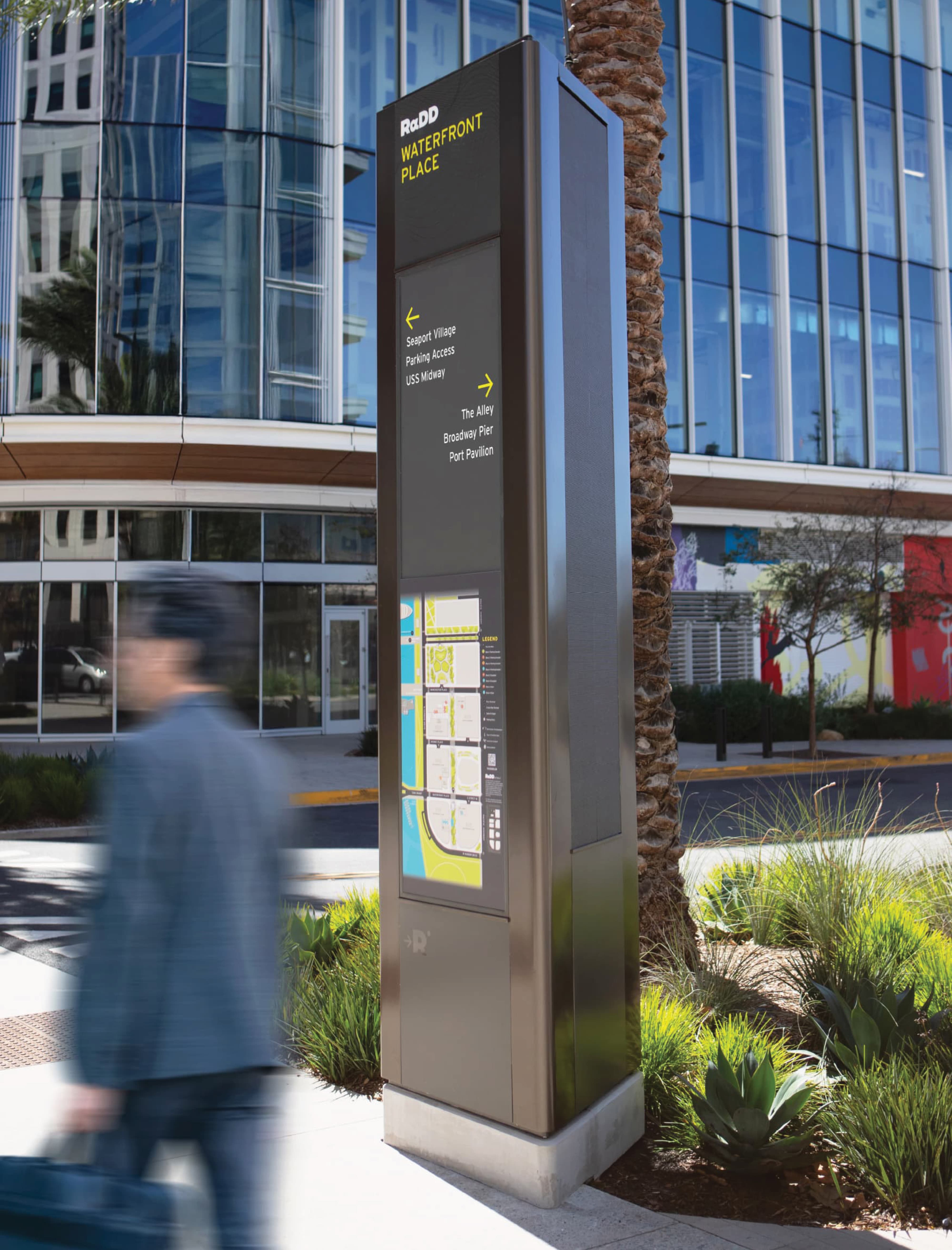 Tall metallic wayfinding pylon reading 'RαDD Waterfront Place,' with directional listings for Seaport Village, Parking Access, USS Midway, The Alley, Broadway Pier, and Port Pavilion, plus an area map below, positioned beside a palm tree in front of a glass office building as a person walks past in motion blur.