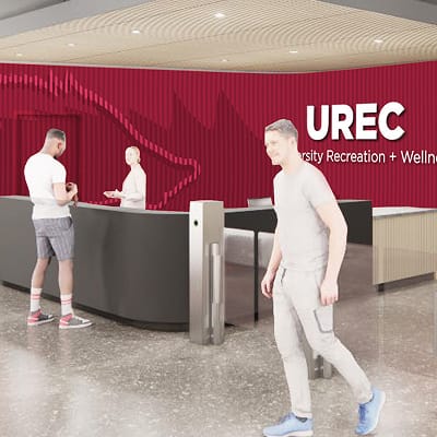 Architectural rendering of the UREC (University Recreation + Wellness) welcome desk. A curved reception counter sits in front of a bold red textured accent wall displaying the "UREC University Recreation + Wellness" logo. A staff member stands behind the desk assisting a visitor, while another person walks through a turnstile entry gate in the foreground. The space features polished floors and a wood-paneled ceiling.
