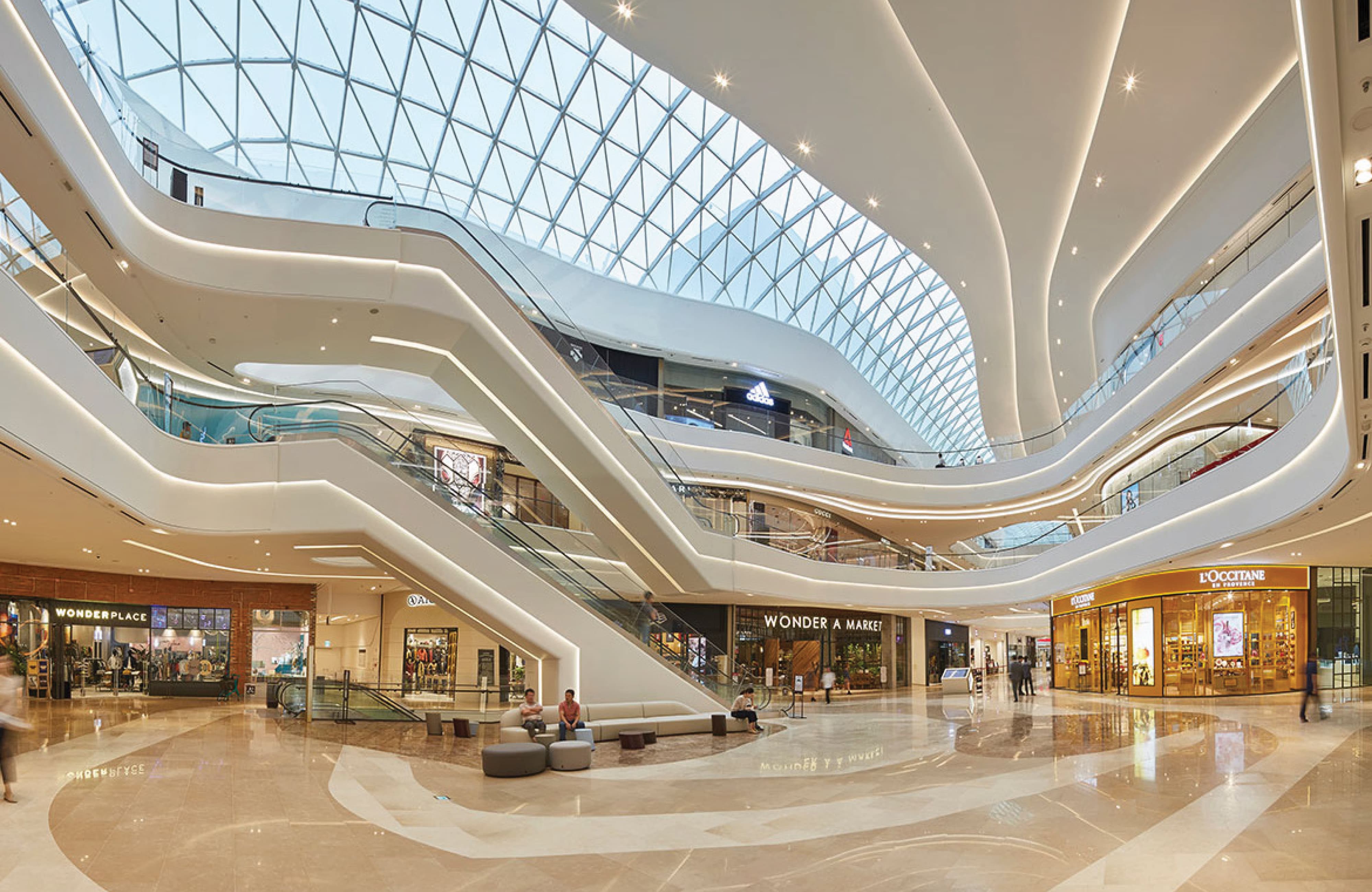 Multi-level shopping mall atrium with sweeping curved white balconies and escalators beneath a triangulated glass skylight, storefronts including L'Occitane, Wonder A Market, and Wonderplace visible below, with shoppers walking across the polished floor.