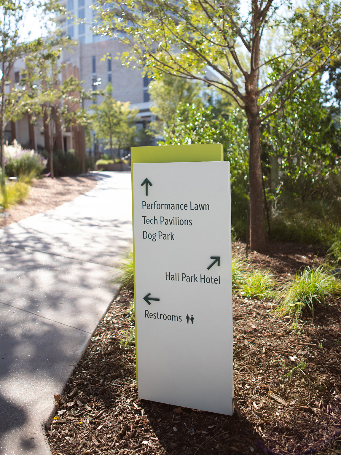 Kaleidoscope Park white and lime green outdoor wayfinding sign with directional arrows pointing to Performance Lawn, Tech Pavilions, Dog Park, Hall Park Hotel, and Restrooms along tree-lined pedestrian path, Frisco, Texas