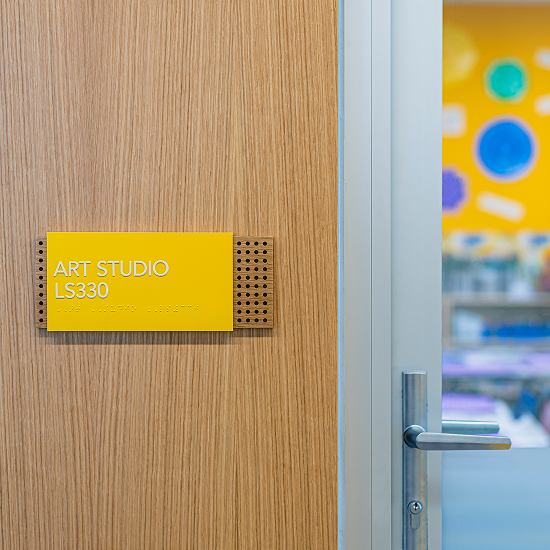 Yellow room sign reading 'Art Studio LS330' with braille, mounted on a wood door beside a glass door revealing a colorful classroom with circular decorations.