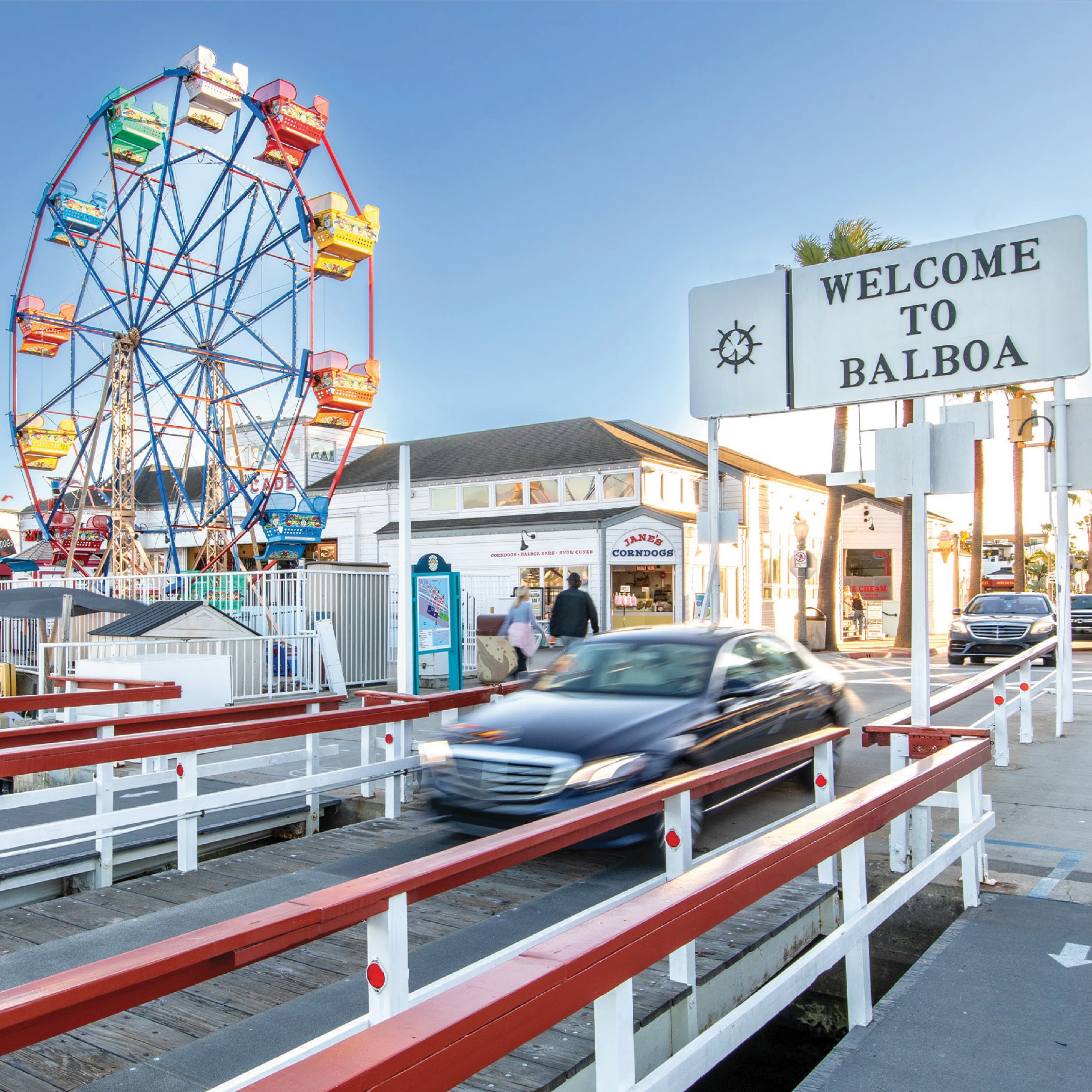 A street-level view of the Balboa Fun Zone waterfront district featuring a "Welcome to Balboa" identity sign with a ship's wheel motif, a colorful Ferris wheel, Jane's Corndogs food and beverage tenant signage, a teal wayfinding kiosk, red and white ferry dock railing, and passing vehicular traffic under palm trees — demonstrating RSM Design's identity signage, wayfinding and signage design, and placemaking services applied within the waterfront, retail, and food and beverage markets