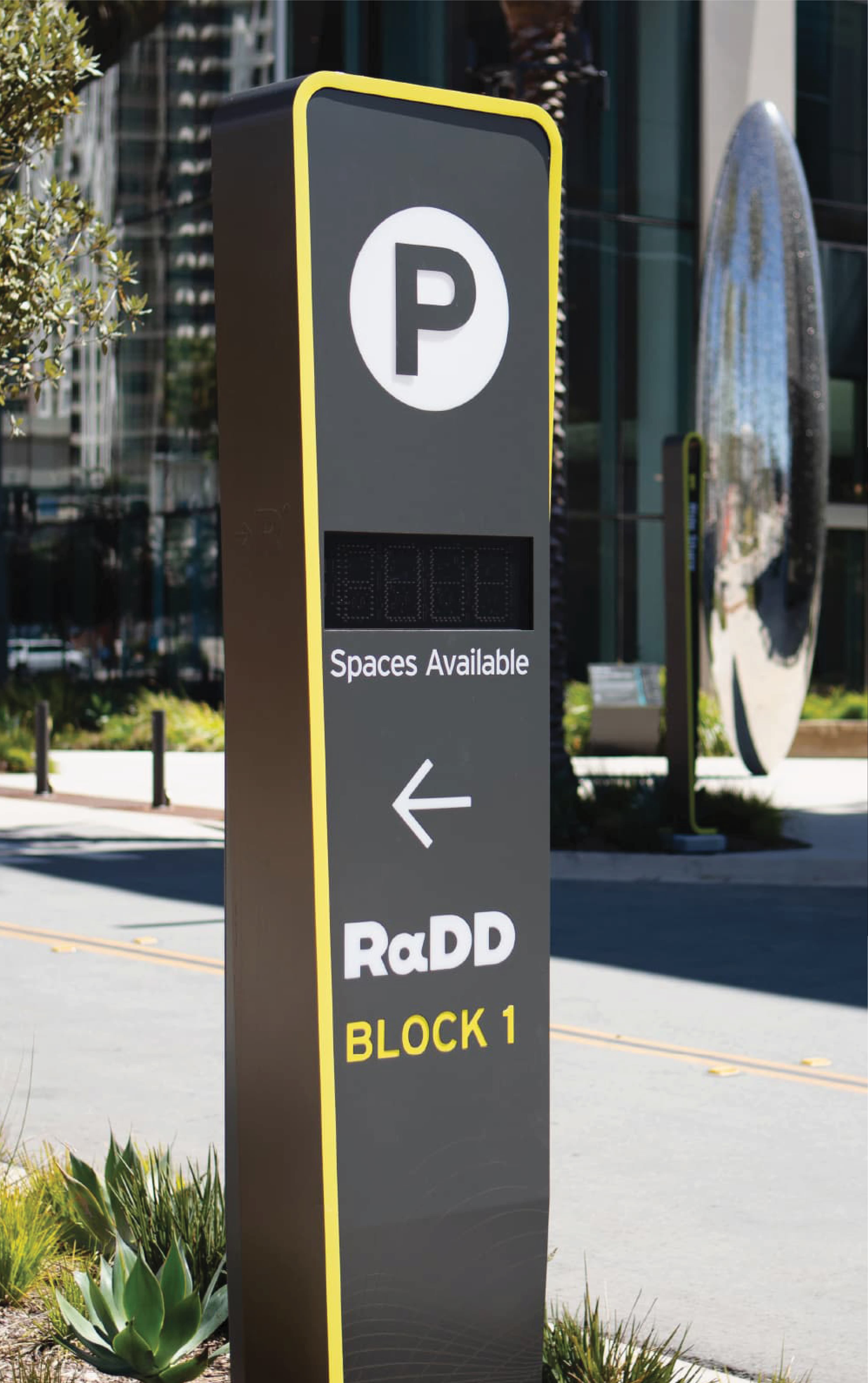 Black parking pylon sign with yellow trim showing a 'P' icon, a digital 'Spaces Available' display, a left-pointing arrow, and 'RαDD Block 1' lettering, positioned near a street with a modern building and reflective sculpture in the background.