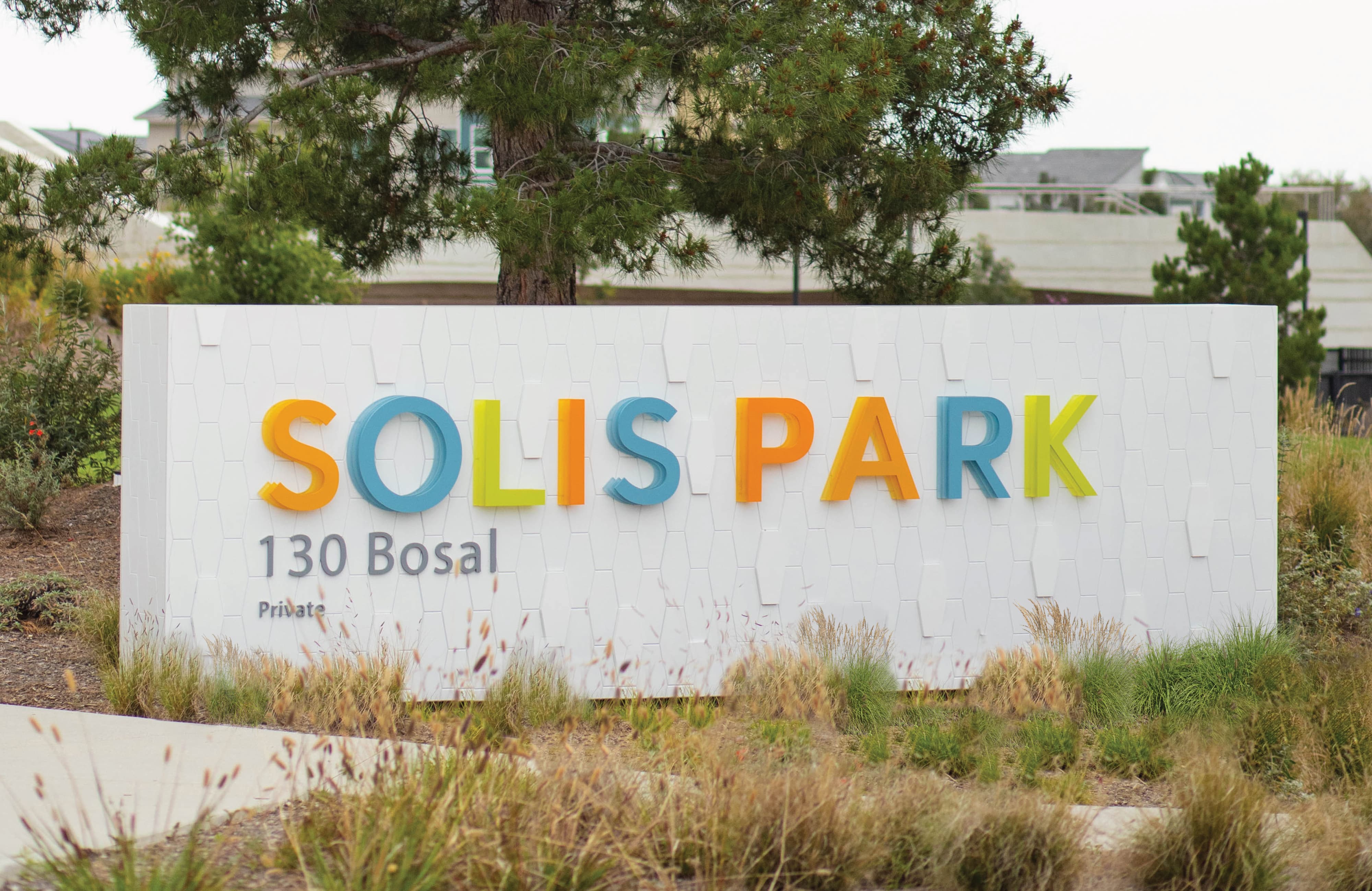 Exterior community monument sign for Solis Park at 130 Bosal featuring large-scale dimensional letters in alternating yellow, teal, orange, and lime green mounted on a textured white tile wall, set among native ornamental grasses and mature pine trees — showcasing RSM Design's identity signage, wayfinding and signage design, and placemaking services within the residential community market