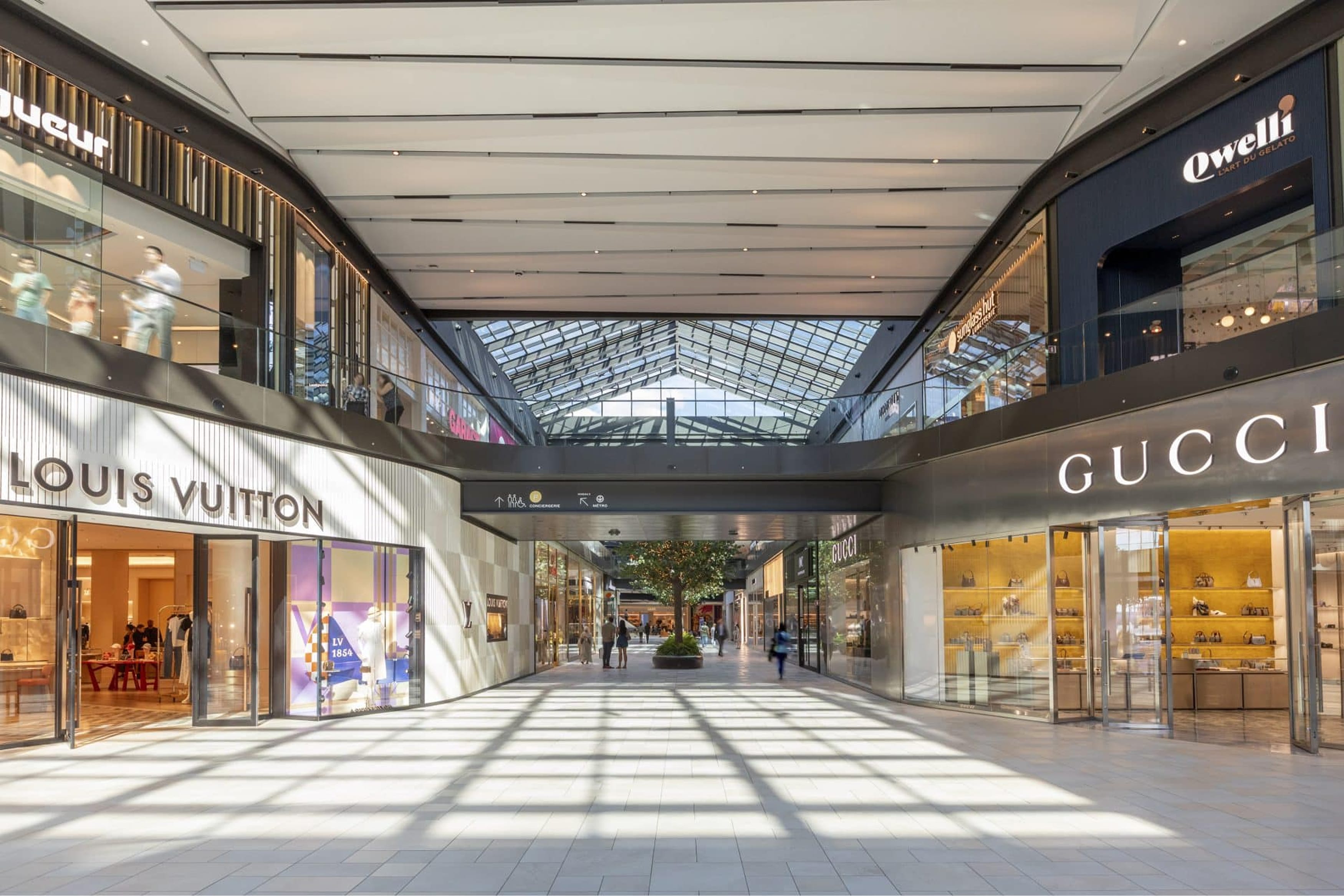Skylit retail concourse at Royalmount with Louis Vuitton and Gucci storefronts, glass roof, and overhead wayfinding sign for concierge and métro — wayfinding design by RSM Design