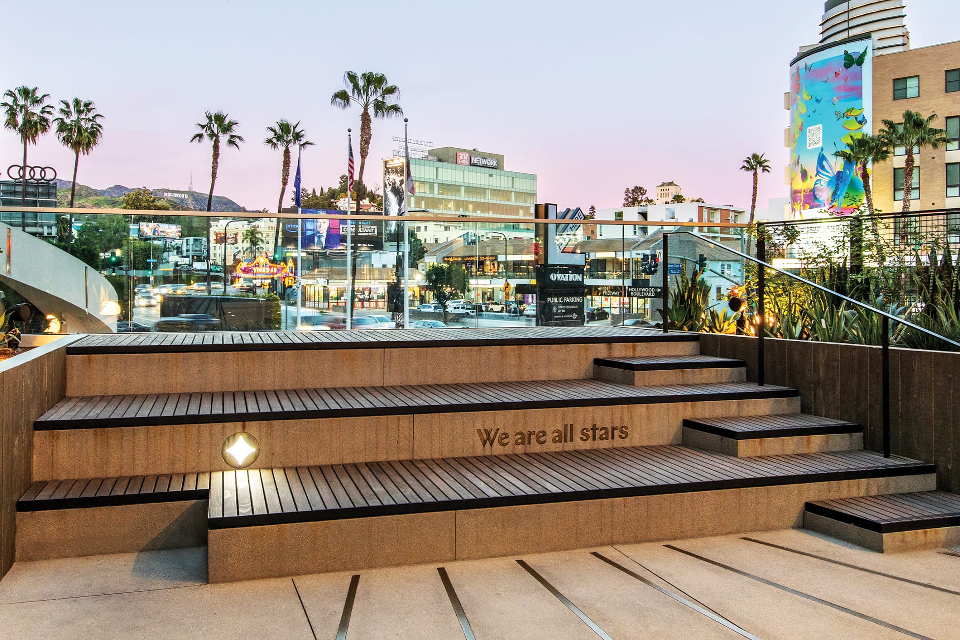 Ovation Hollywood rooftop terrace amphitheater steps with engraved "We are all stars" inscription, illuminated Ovation diamond logo, glass railing overlooking Hollywood Boulevard at dusk with Hollywood Sign visible in background