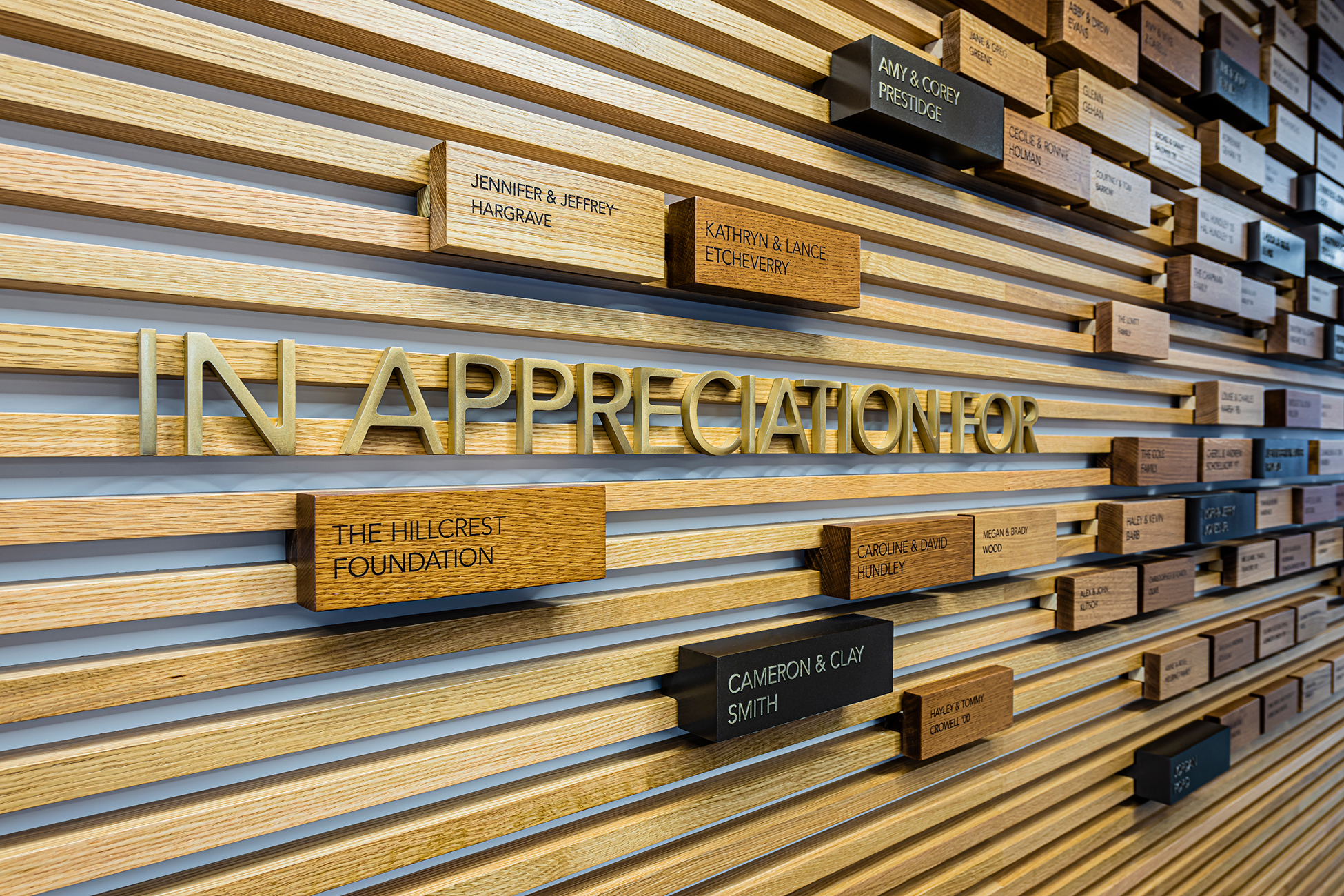 Wood slat donor recognition wall reading 'In Appreciation For' in bronze dimensional letters, with protruding wood and black blocks engraved with donor names such as The Hillcrest Foundation, Jennifer & Jeffrey Hargrave, and Cameron & Clay Smith.