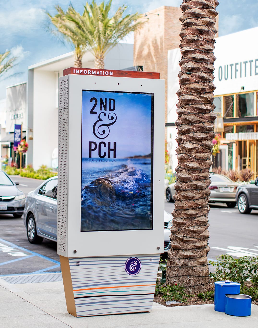 Branded outdoor digital information kiosk at 2nd & PCH in Long Beach, California, displaying the 2nd & PCH logo over a coastal wave photograph on an interactive touchscreen, with a striped base in the brand color palette and an ampersand medallion mark — showcasing RSM Design's digital integration and interactive solutions, wayfinding signage design, and branded environments for a coastal waterfront retail and mixed-use destination.