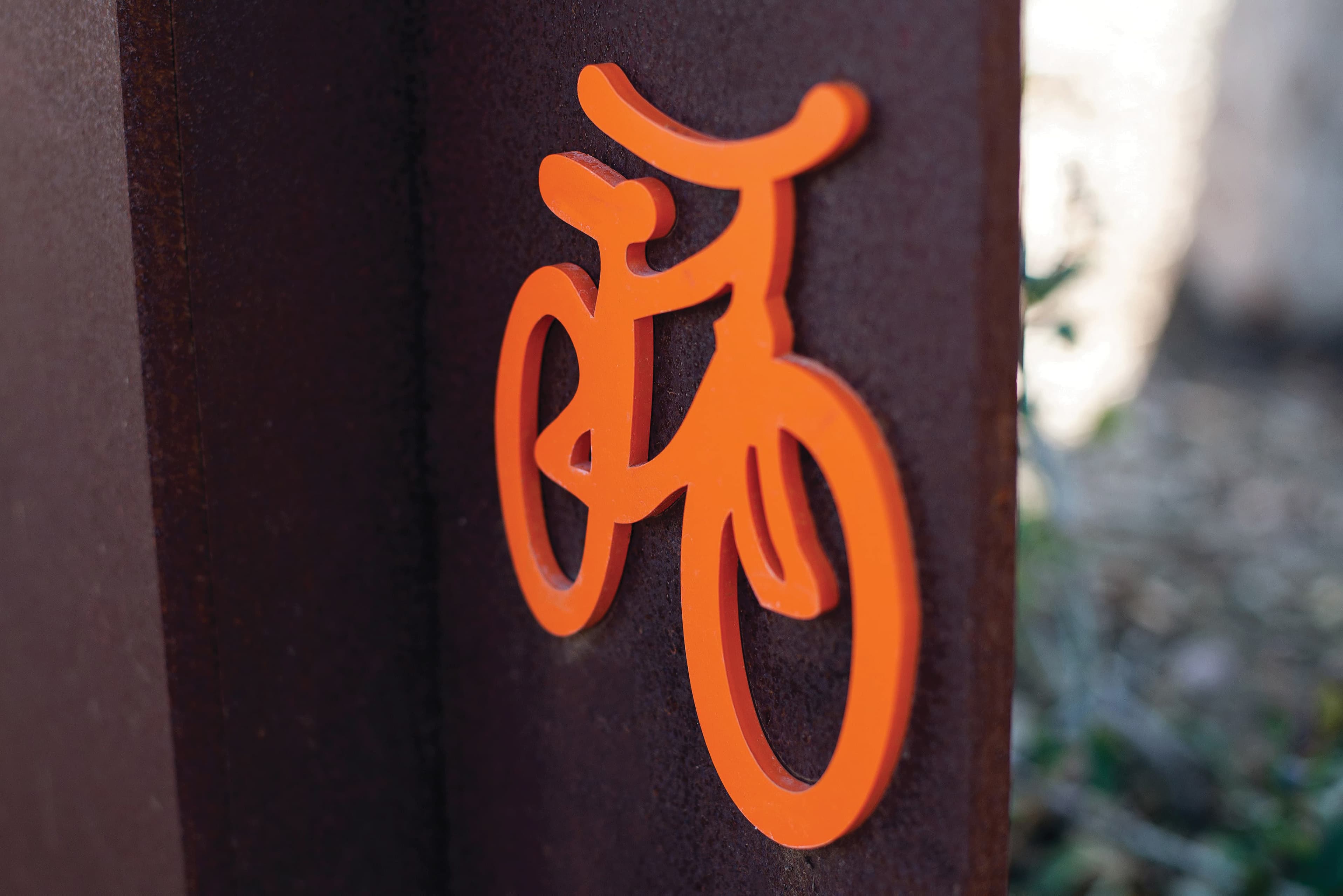 Close-up of an orange dimensional bicycle icon mounted on a rusted corten steel panel.
