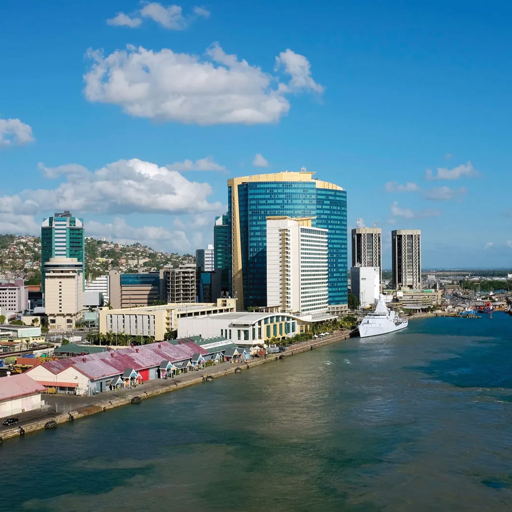 Panoramic waterfront skyline view of Port of Spain, Trinidad and Tobago, showing modern high-rise office towers, waterfront buildings, and docks along the Caribbean coast.