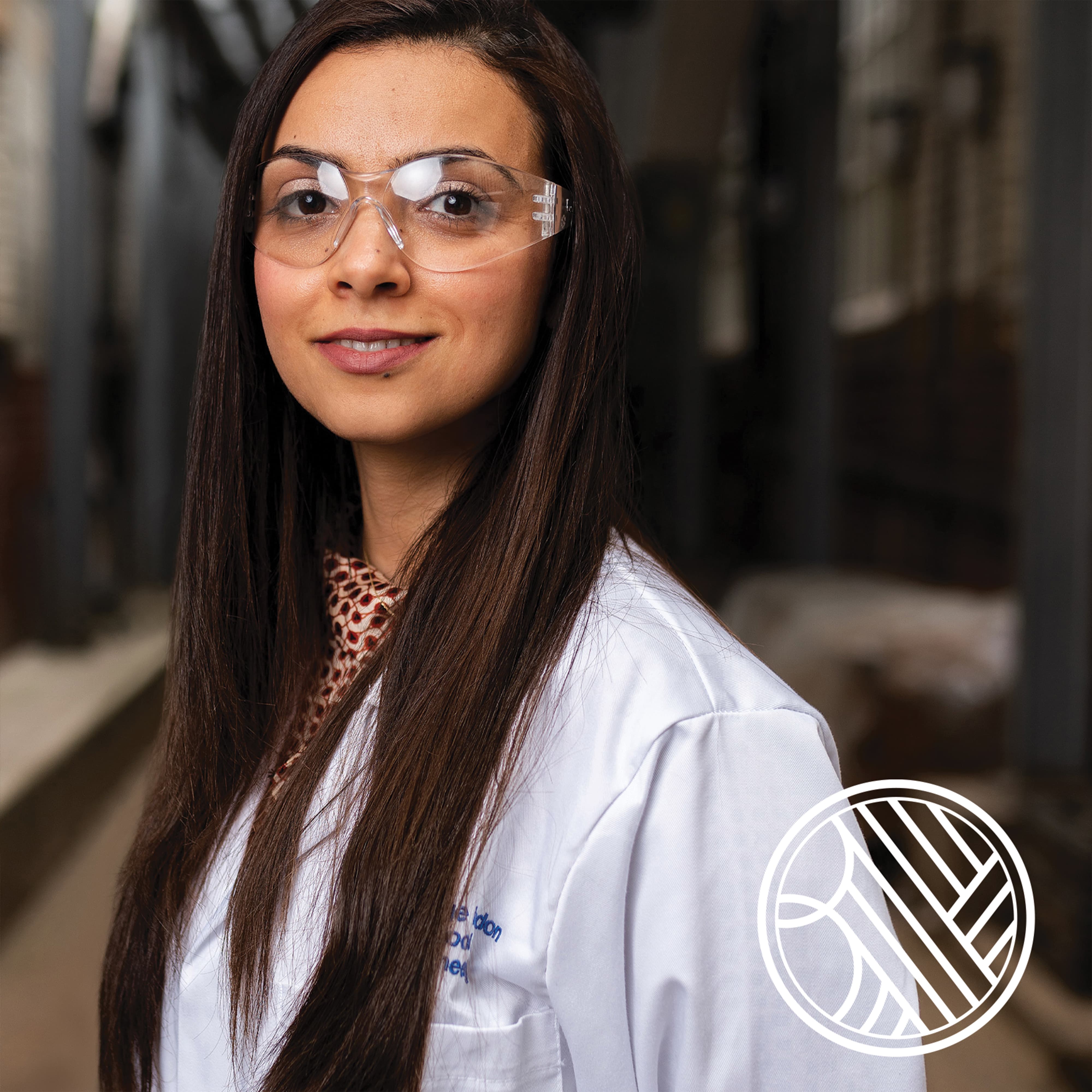 Portrait of a woman wearing safety glasses and a white lab coat, smiling at the camera in an industrial setting, with the Biovista logo mark overlaid in the bottom right corner.