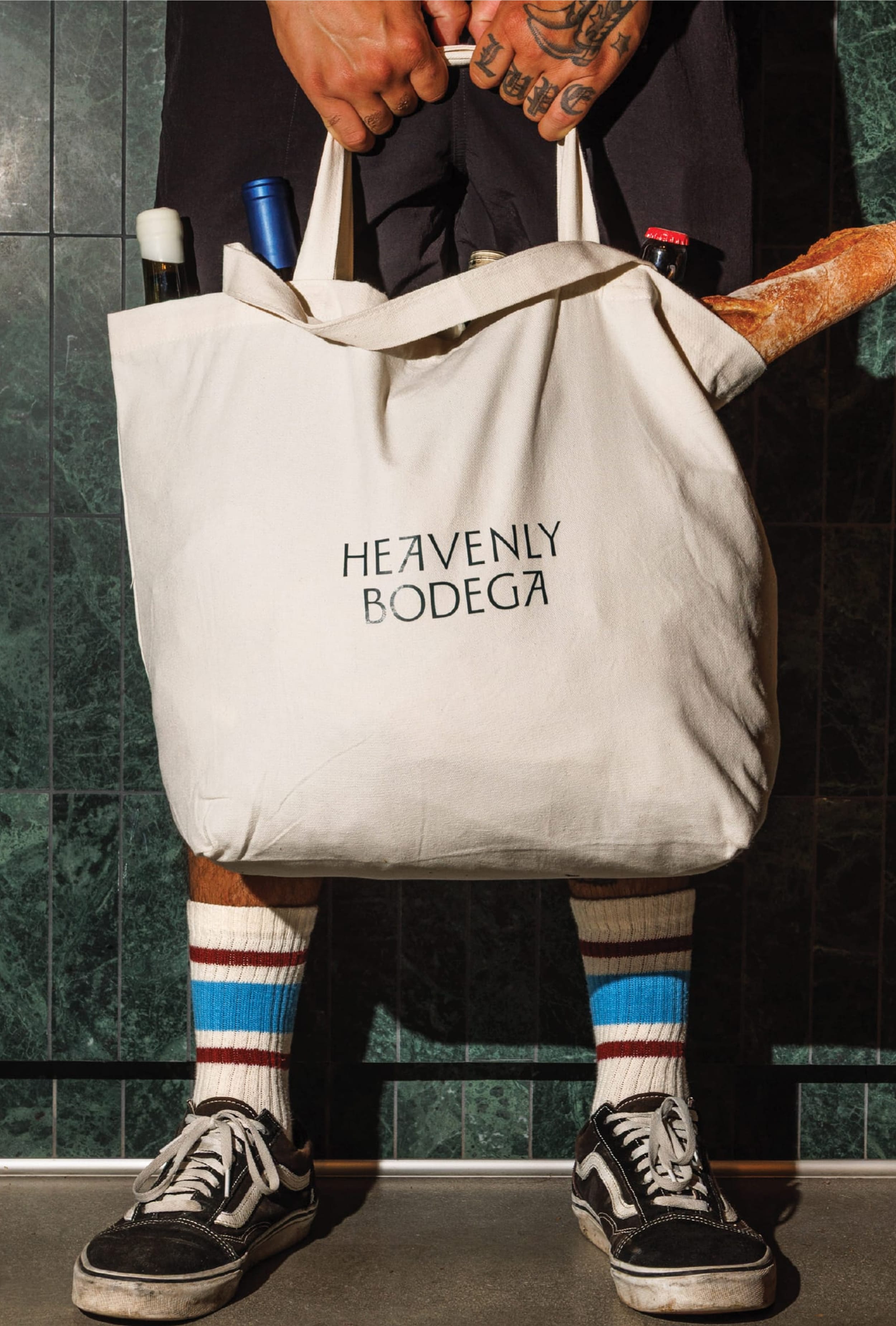 Person holding a canvas "Heavenly Bodega" tote bag loaded with wine bottles and a baguette, standing on green tile flooring — branded merchandise design by RSM Design