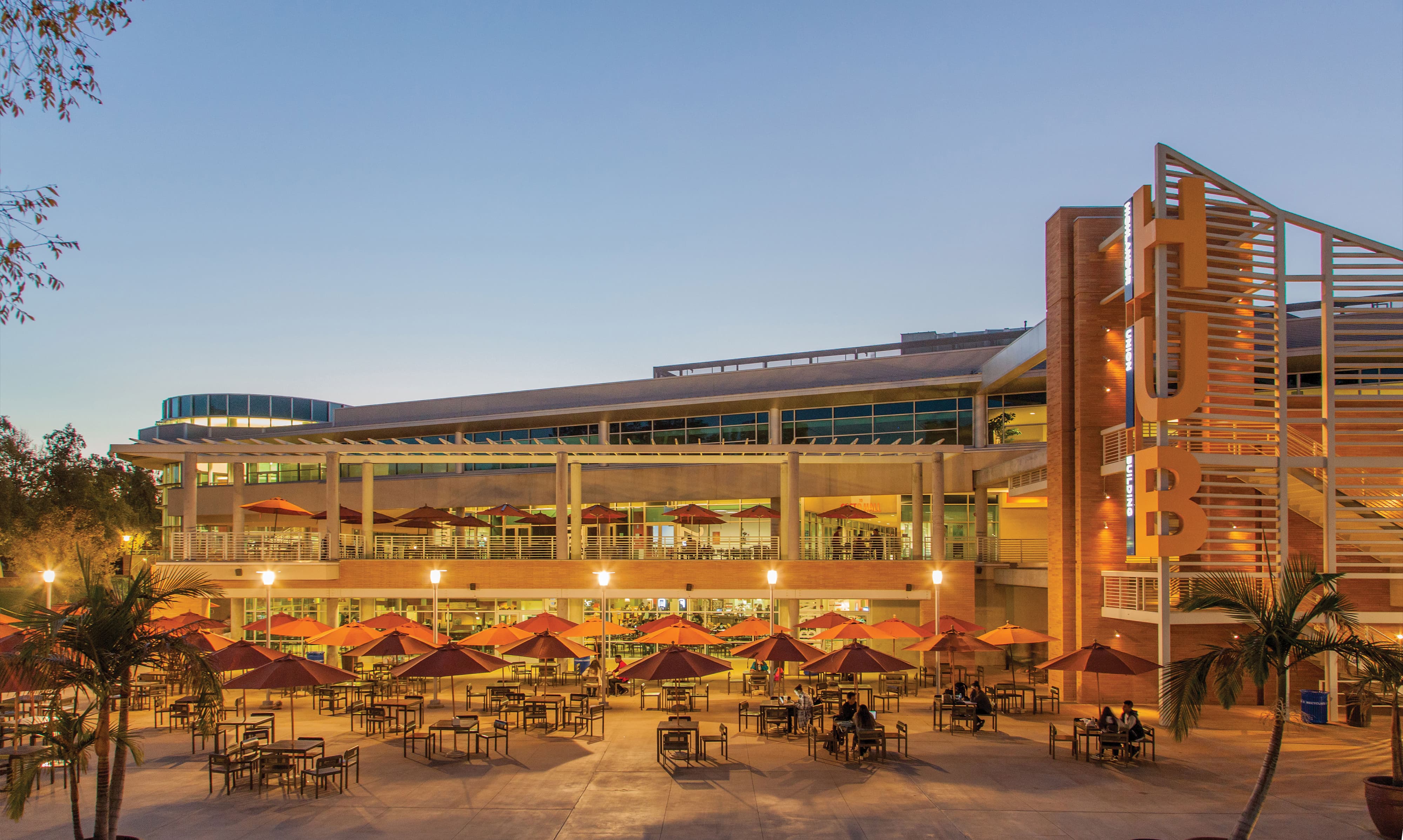 Twilight view of the illuminated Highlander Union Building with a large orange 'HUB' letter sculpture, its outdoor patio filled with orange umbrella-shaded tables where students sit, and palm trees in the foreground.