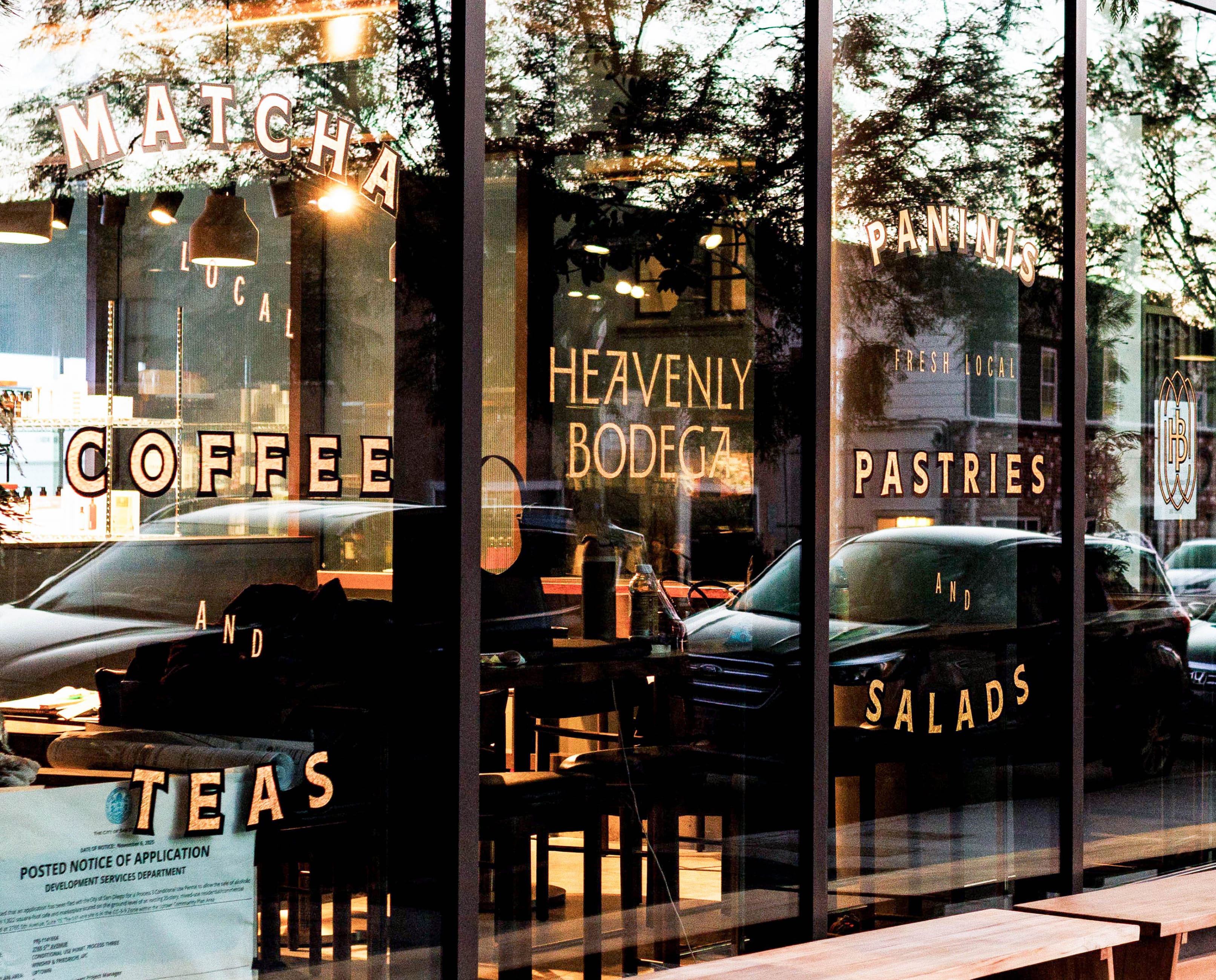 Storefront window lettering for Heavenly Bodega reading "Matcha, Coffee and Teas, Panninis, Fresh Local Pastries and Salads" with tree reflections and street view — window graphics design by RSM Design