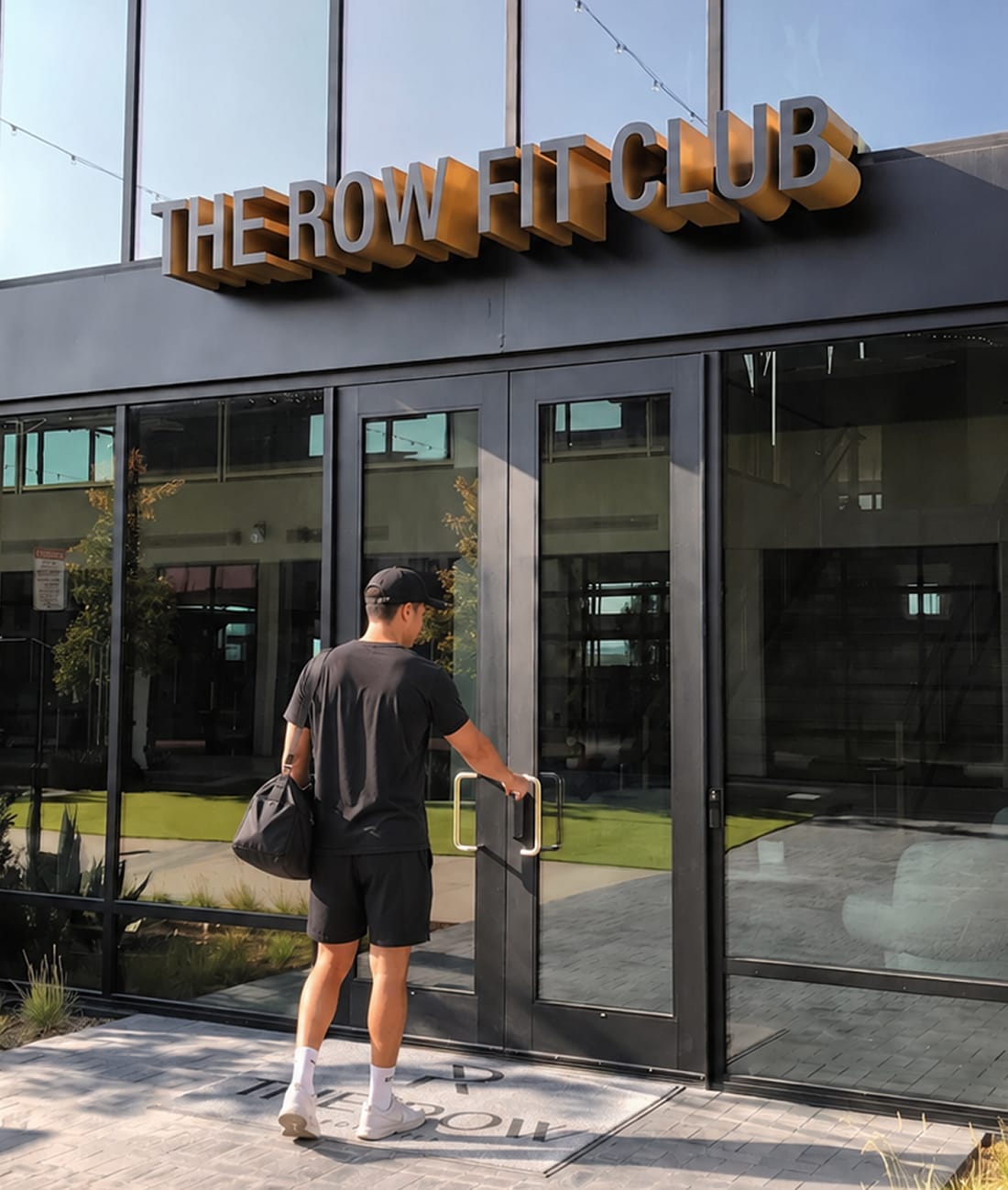 Dimensional bronze "THE ROW FIT CLUB" building sign above black-framed glass entry doors, with a resident entering the fitness club at The Row at Red Hill – placemaking and environmental graphic design by RSM Design.