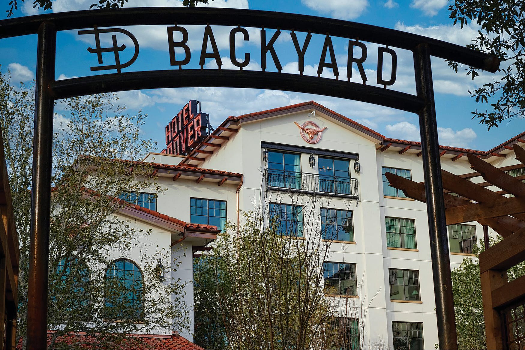Wrought iron Backyard entrance gate at Hotel Drover in Fort Worth Stockyards, Texas, framing the hotel's stucco facade, longhorn emblem, red tile roof, and rooftop neon sign against a blue sky