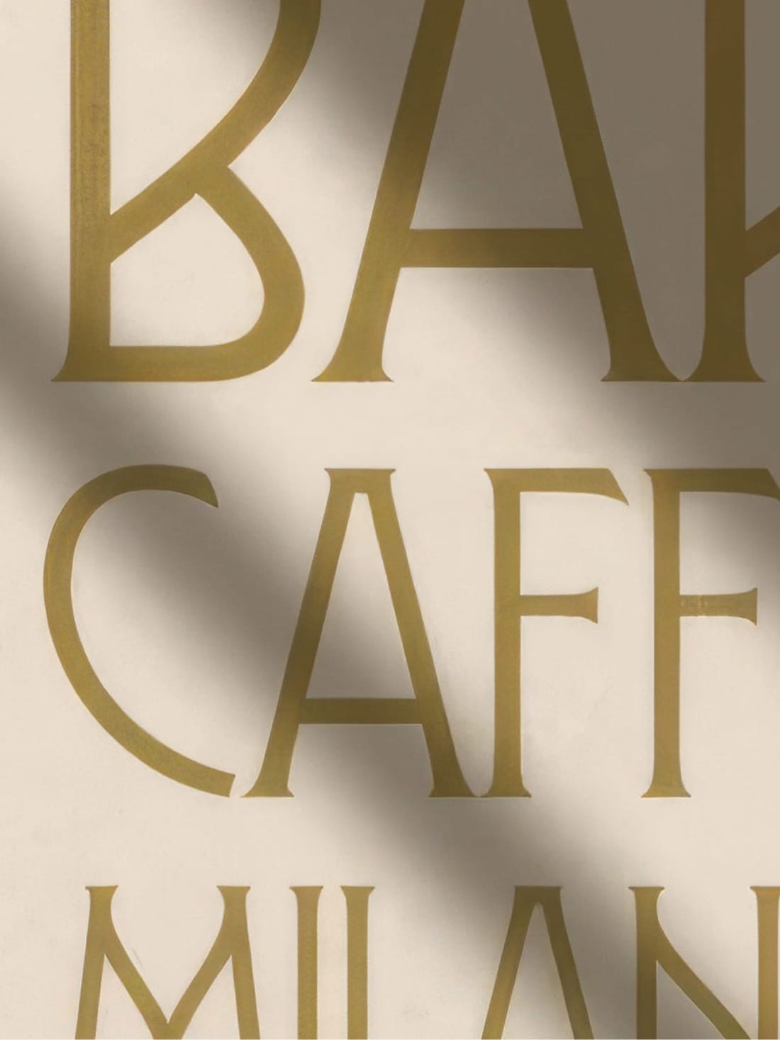 Close-up of gold serif lettering reading "Bar," "Caffè," and "Milan" with soft cast shadow across cream wall — custom typography and signage by RSM Design