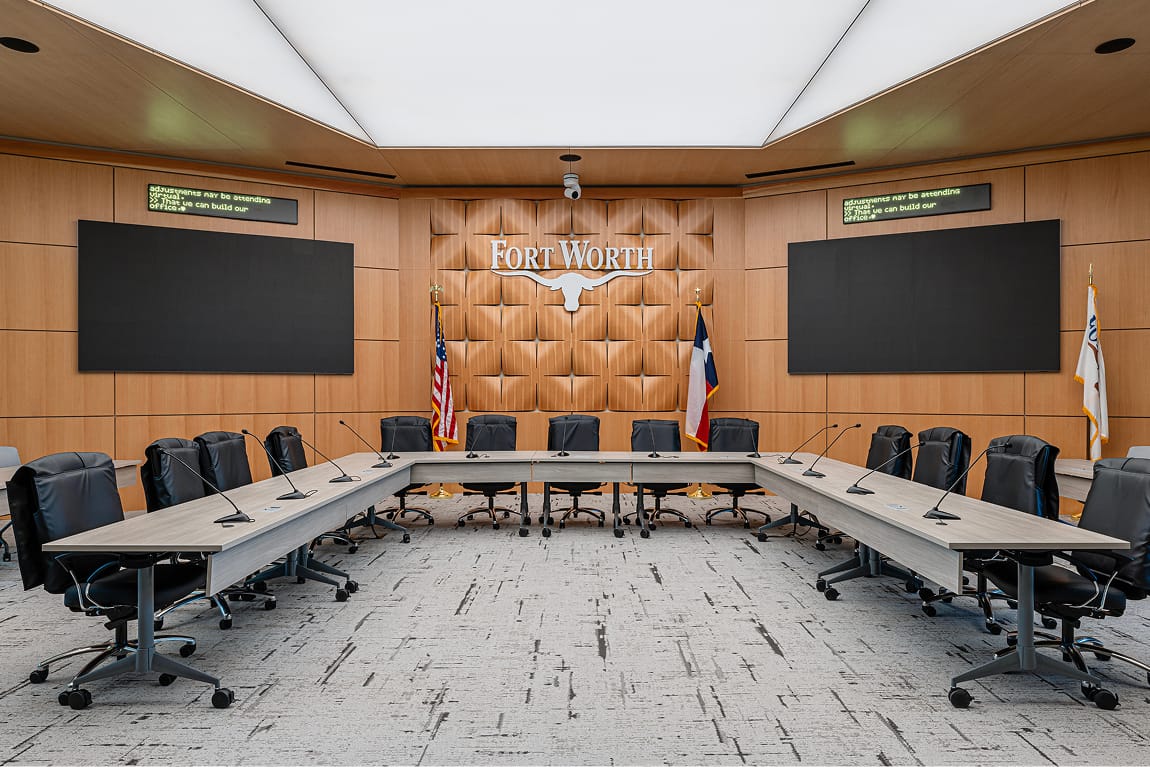 Fort Worth City Hall conference room interior featuring dimensional Fort Worth longhorn logo on sculpted wood panel wall, U-shaped conference table with microphones, dual display screens, and Texas and American flags